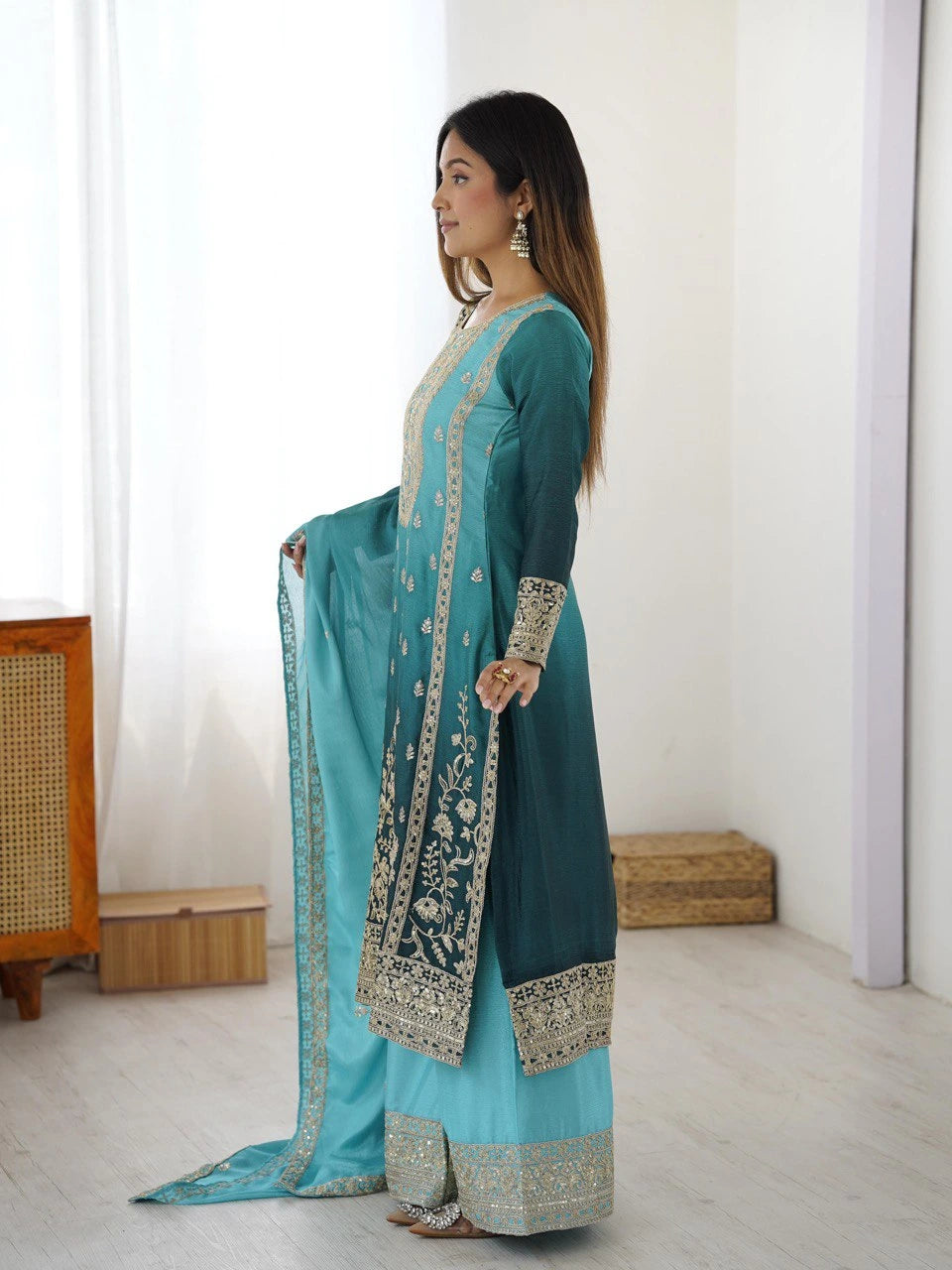 Woman wearing a teal and light blue traditional outfit with embroidery in a minimalistic room.