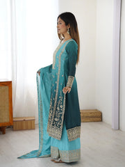 Woman wearing a teal and light blue traditional outfit with embroidery in a minimalistic room.