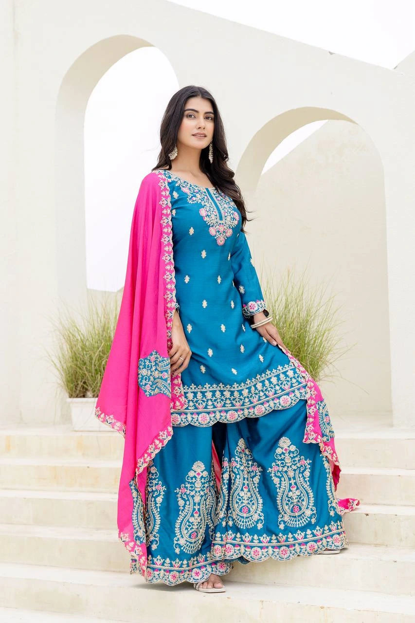 Woman in a blue embroidered traditional outfit with pink dupatta standing on steps.