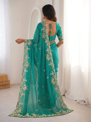 Woman wearing a teal saree with floral patterns in a bright room.