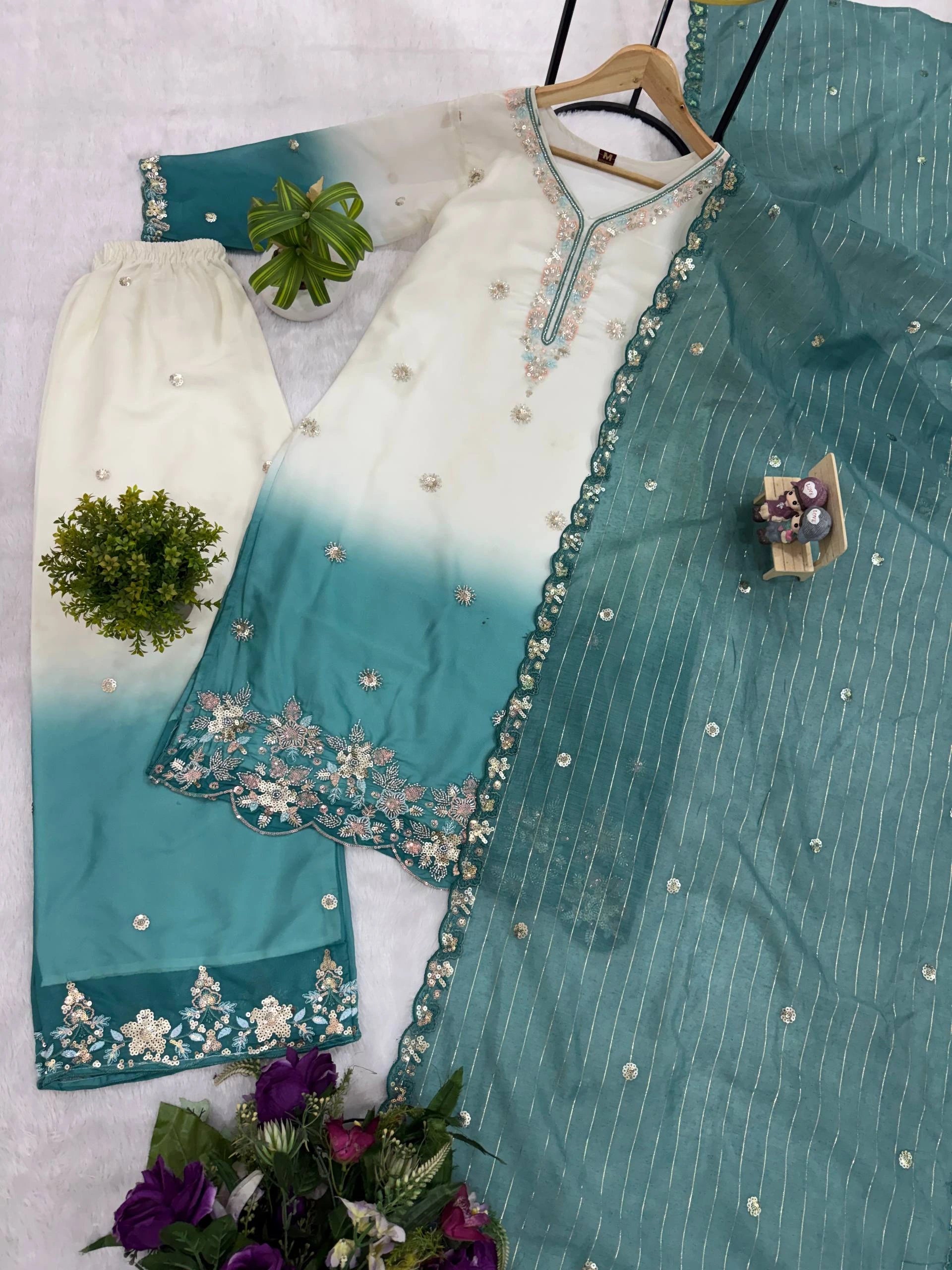 Teal and white embroidered traditional outfit on a light gray background with green plants.