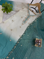 Teal and white embroidered garment on a hanger with a small table and chairs model below.