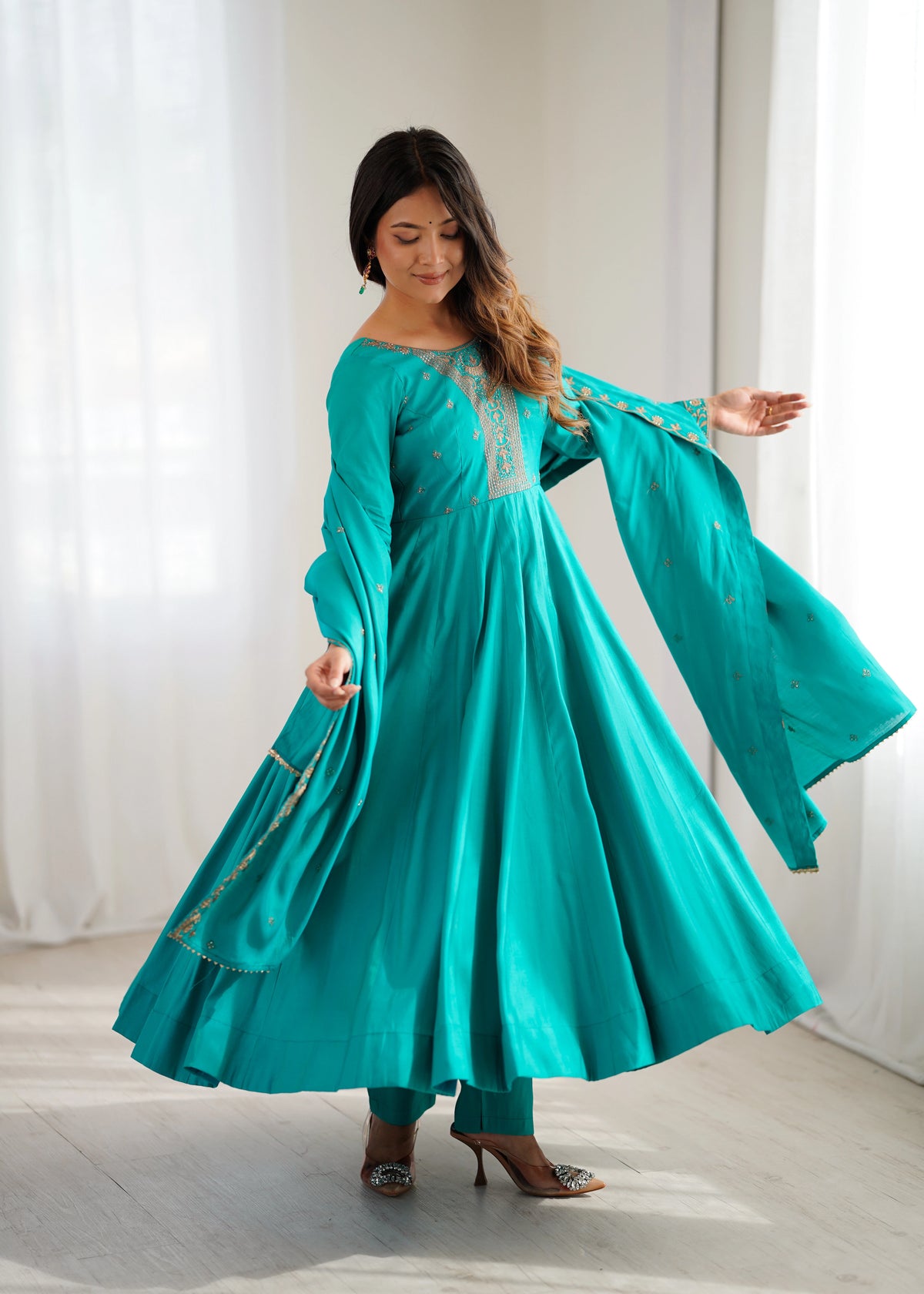 Woman wearing a turquoise traditional outfit with gold accents in a bright room.