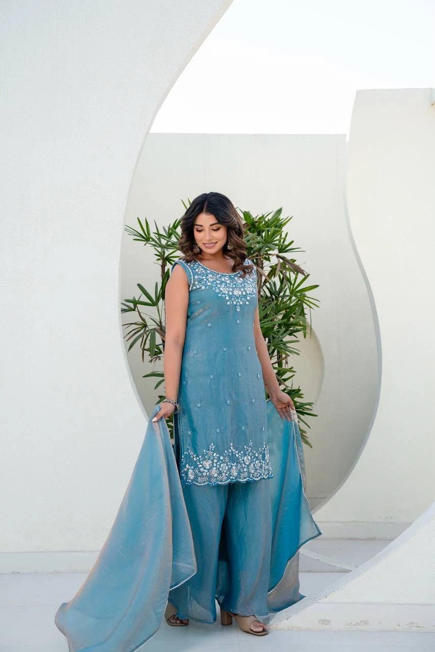 Woman in a light blue embroidered outfit standing against a white background with green plants.