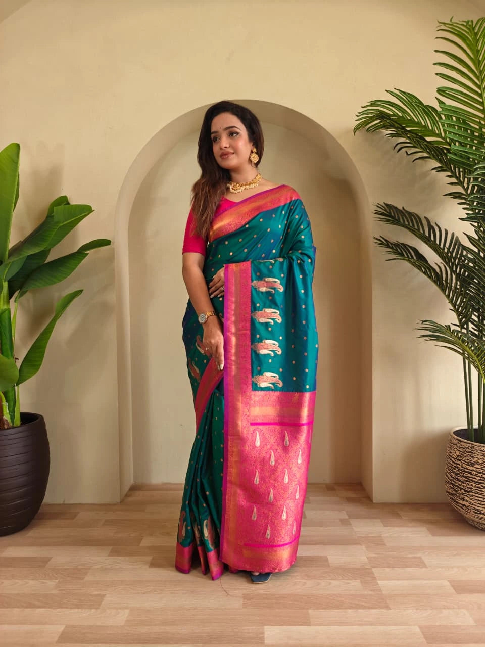 Woman in a teal and pink saree standing in a room with plants.
