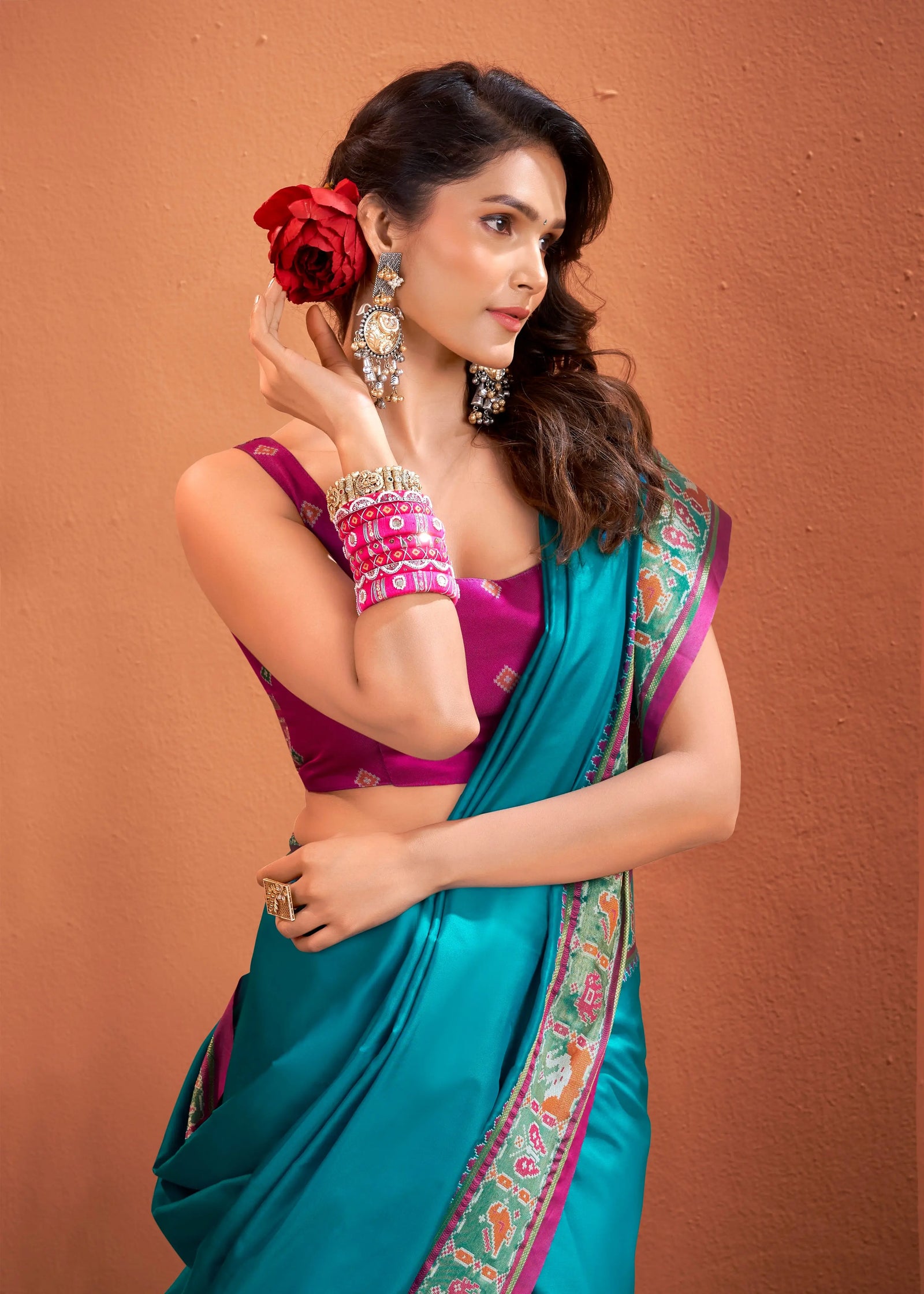Woman wearing a blue saree with a purple blouse, holding a red flower against a brown background