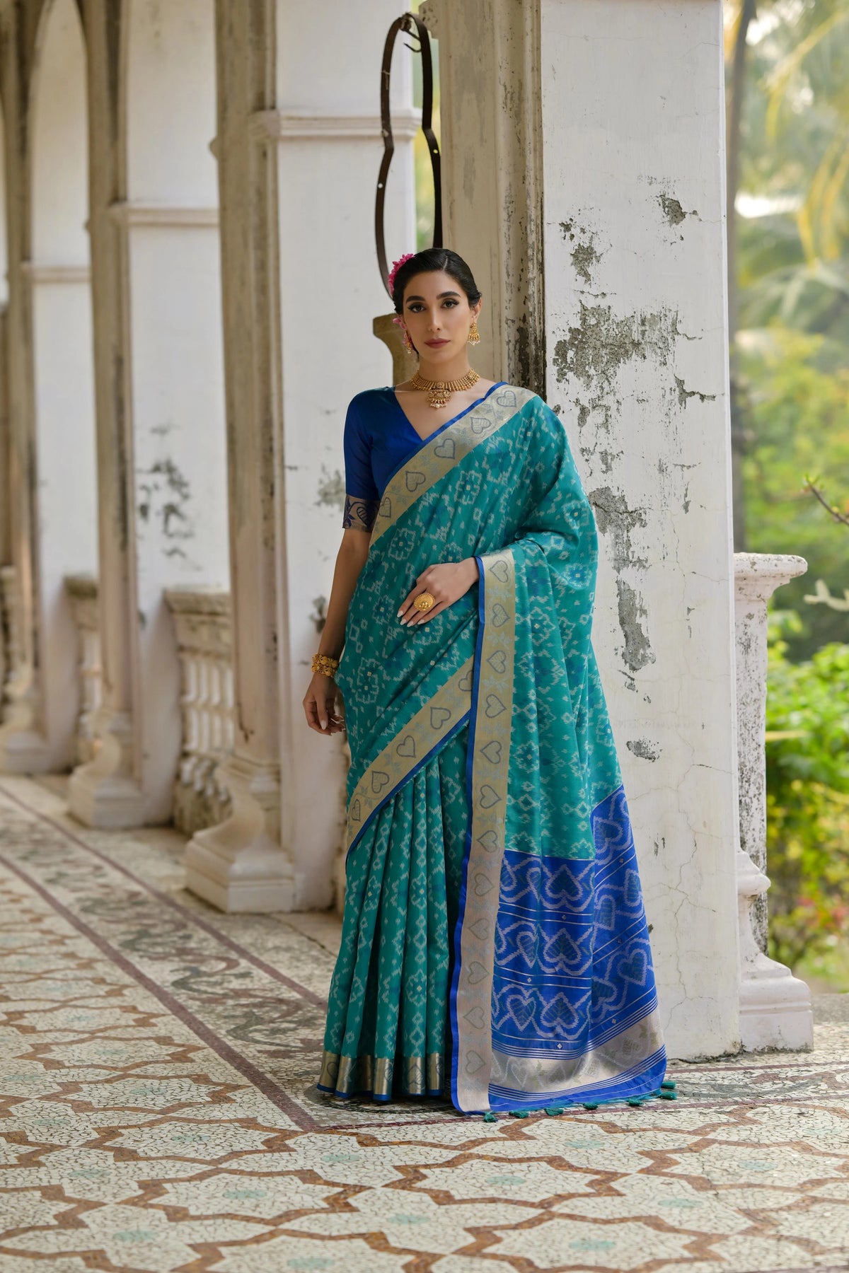 Woman in a blue and green saree standing on a patterned floor with a white wall and greenery in the background.