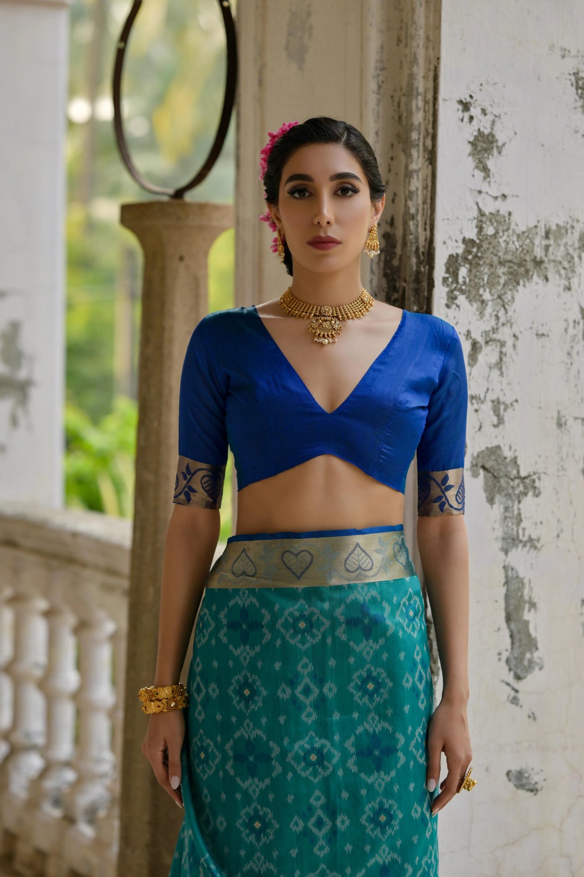 Woman wearing a blue top and green patterned skirt with jewelry in an outdoor setting.