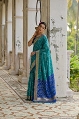 Woman in a teal and blue saree standing in an outdoor setting with classical architecture.