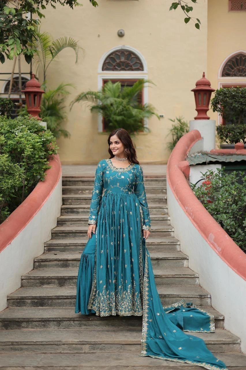 Woman in a teal traditional outfit standing on a staircase with greenery and a building in the background