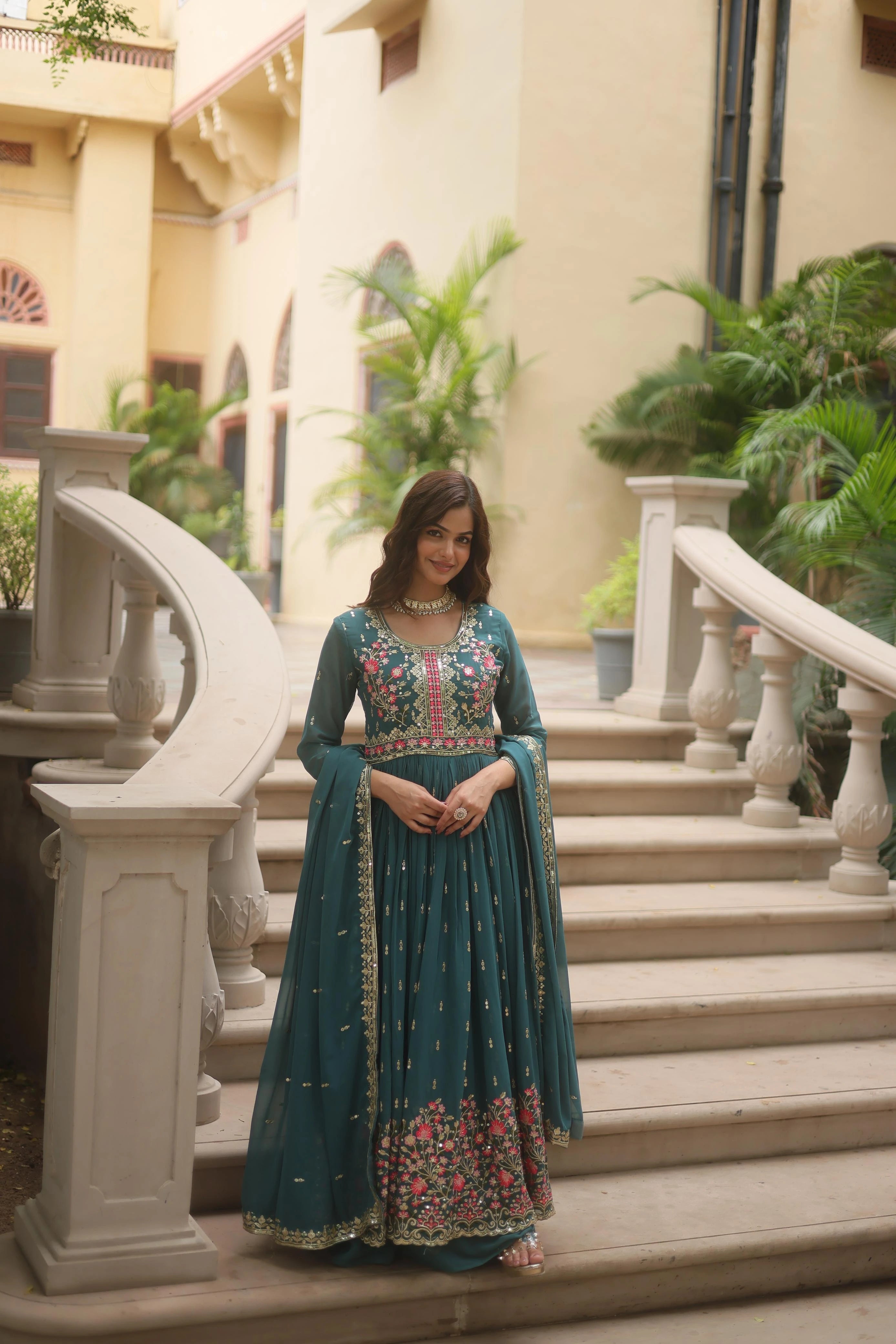 Woman in a teal traditional outfit standing on steps with decorative railings and plants in the background.