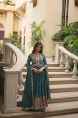Woman in a teal traditional outfit standing on steps with decorative railings and plants in the background.