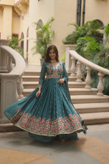 Woman in a teal embroidered dress standing in an elegant indoor setting with plants and stairs.