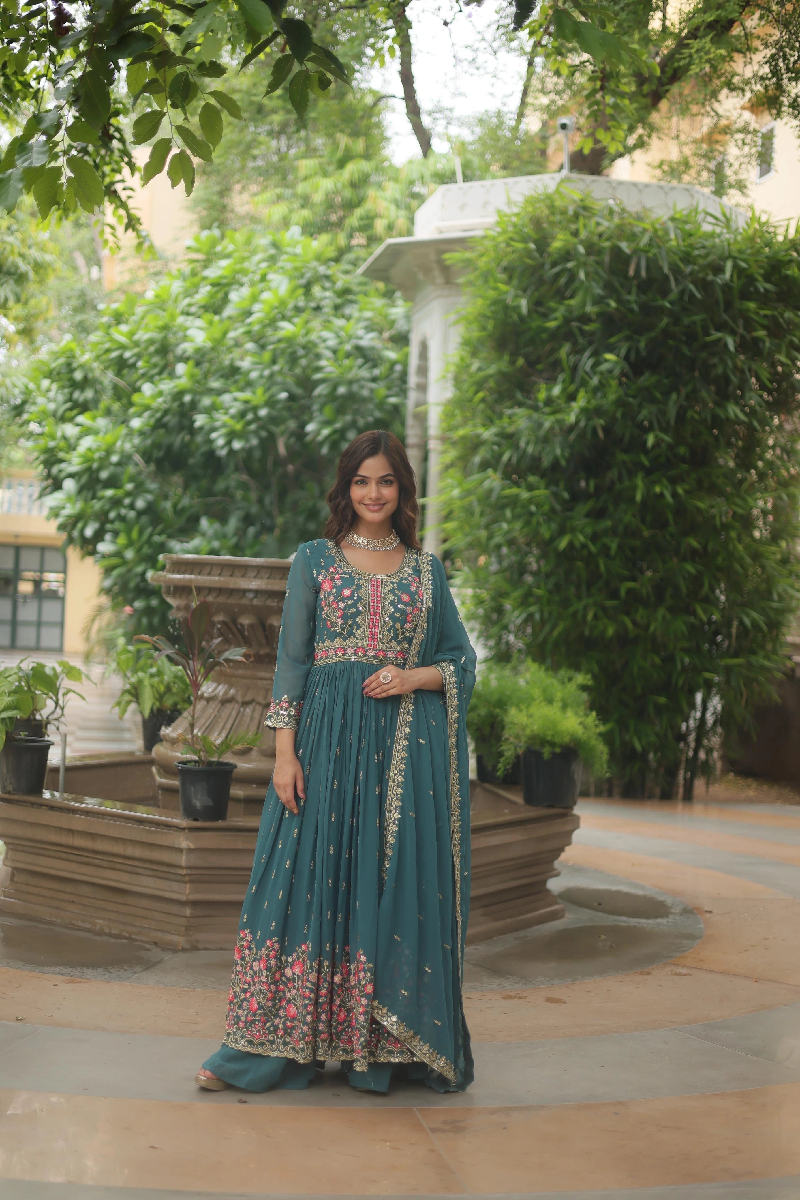 Woman in a teal traditional outfit with floral patterns standing outdoors near greenery.