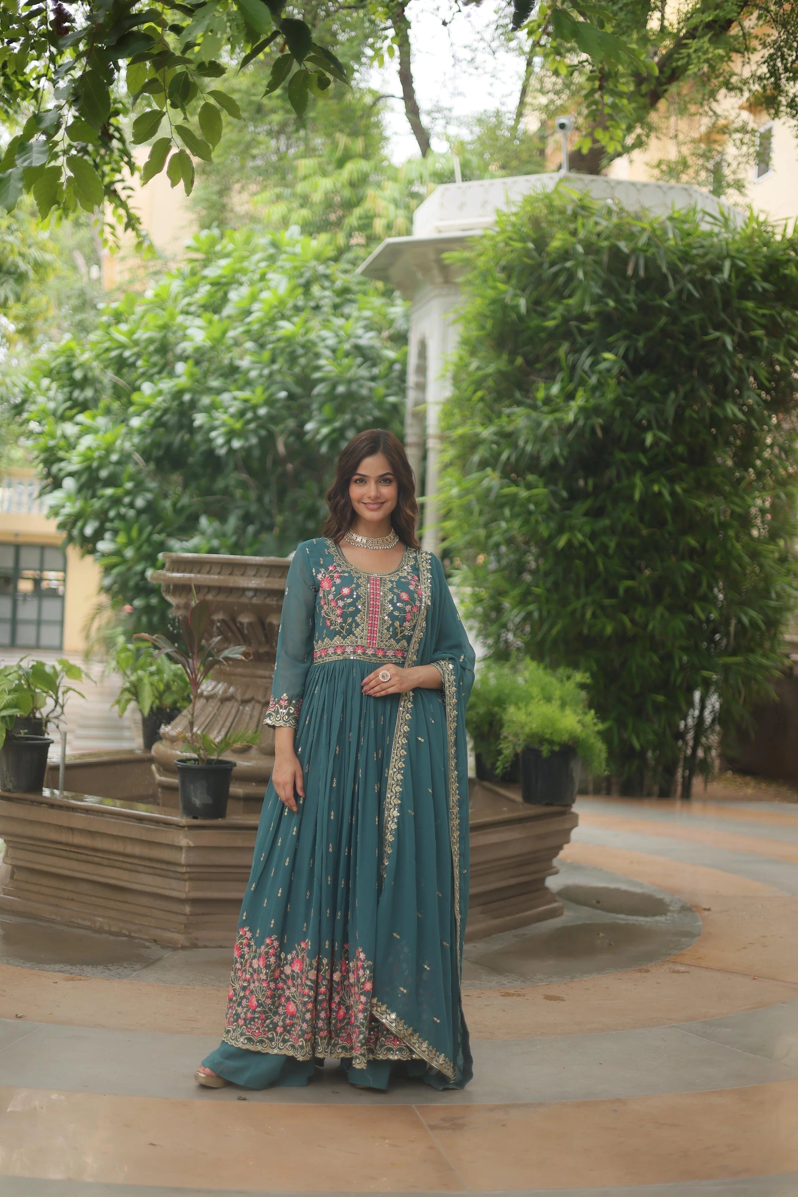 Woman in a teal traditional outfit with floral patterns standing outdoors near greenery.