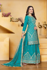 Woman in a teal embroidered traditional outfit standing in a decorative room.