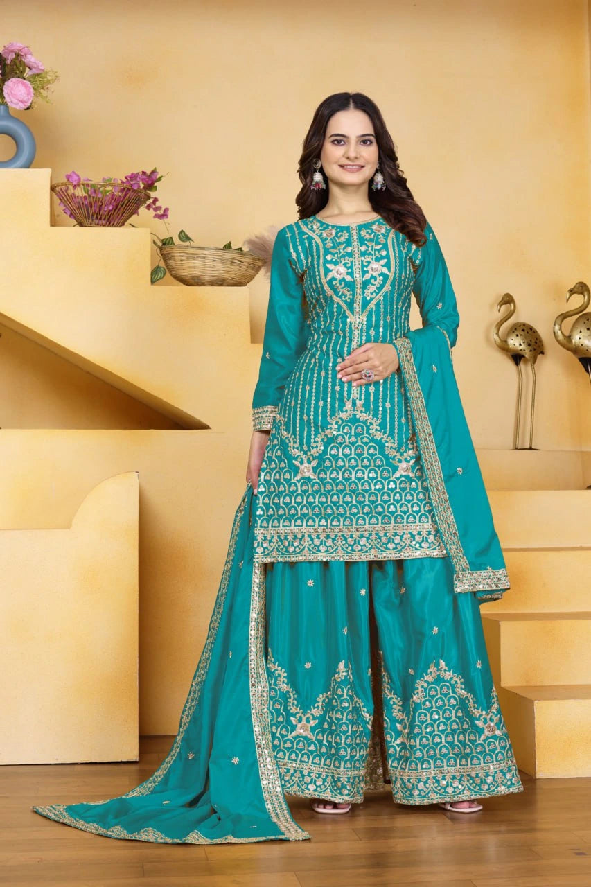 Woman in a teal embroidered traditional outfit standing in a decorative room.