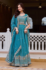 Woman in a teal traditional outfit with intricate patterns standing in a decorative setting.