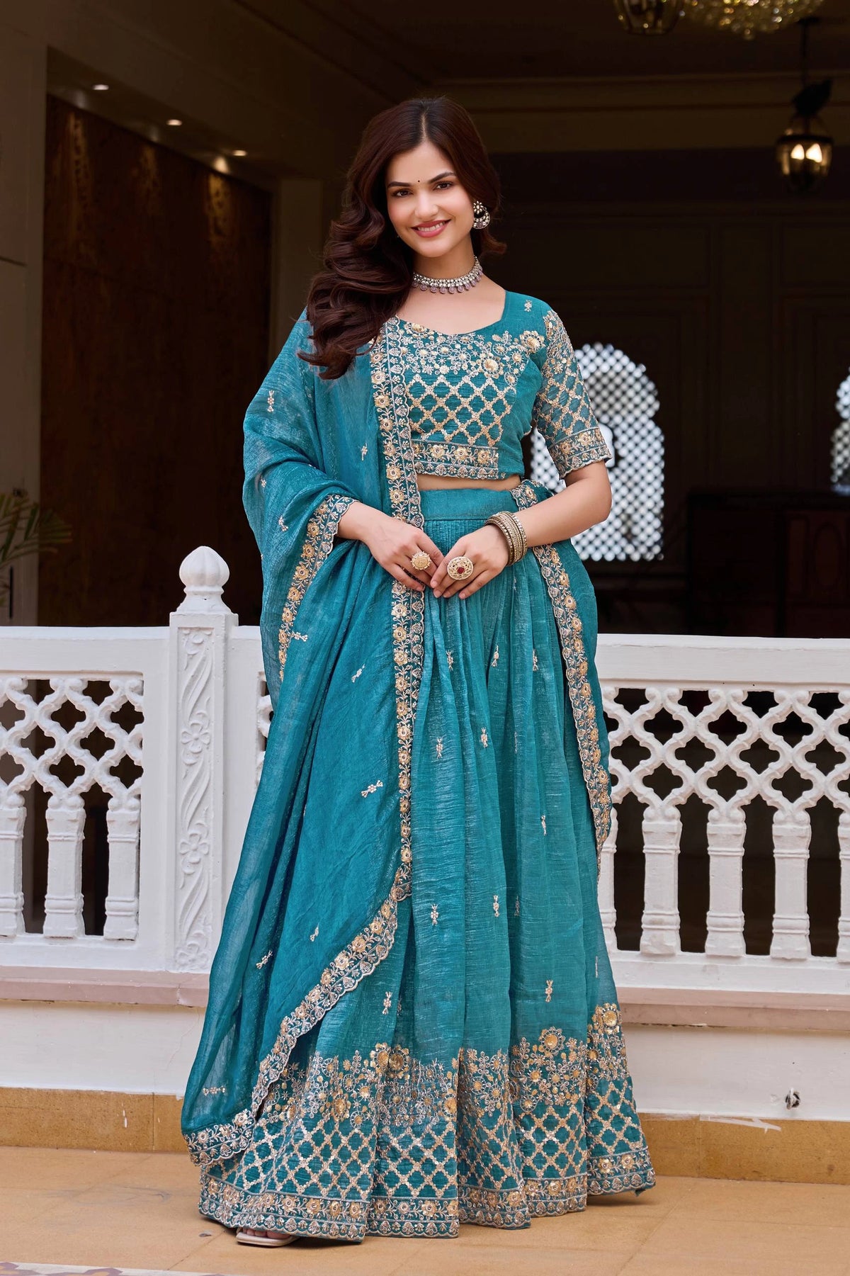 Woman in a teal traditional outfit with gold embroidery standing indoors.