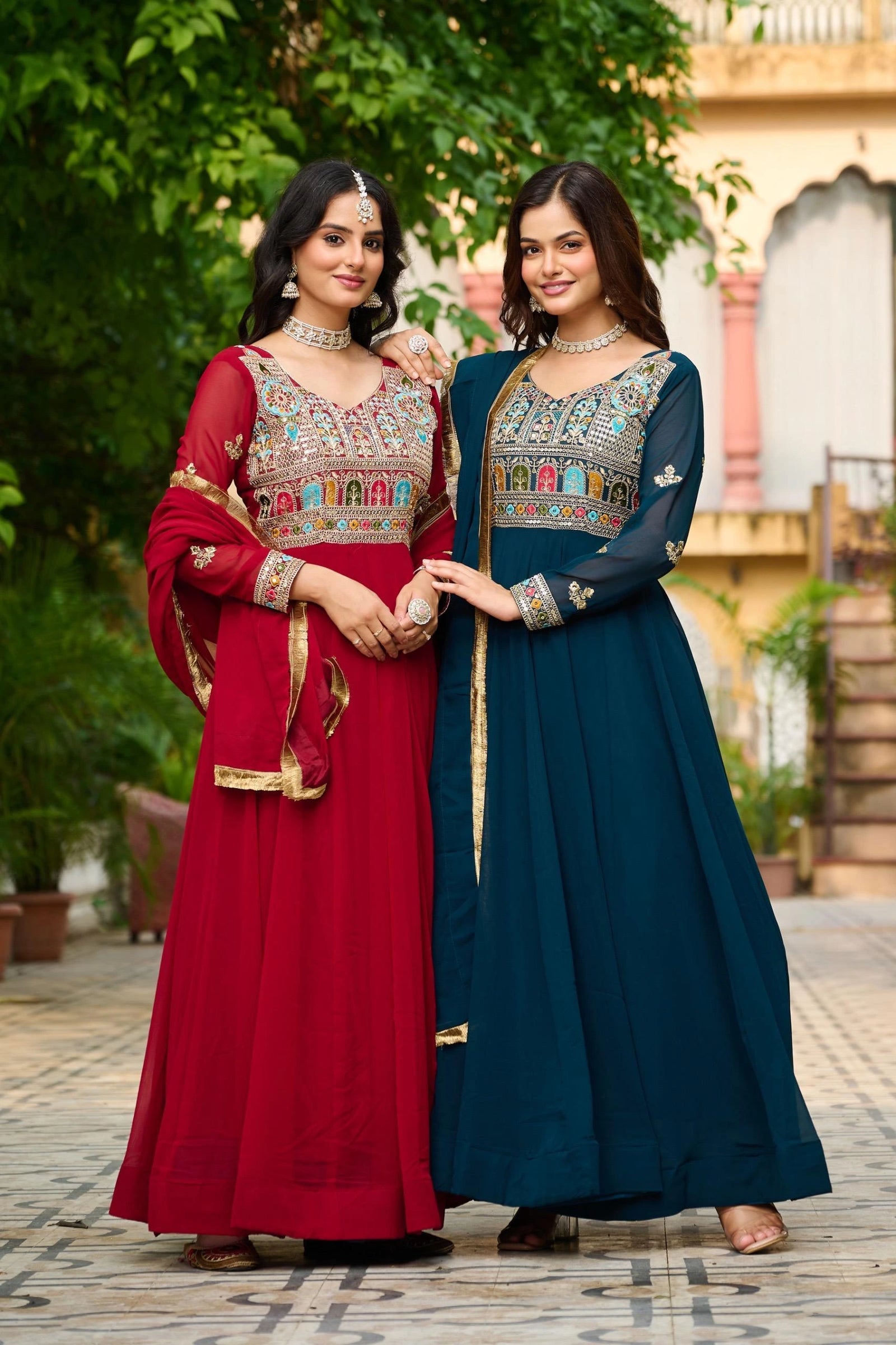 Two women in traditional outfits standing outdoors with a building and greenery in the background.