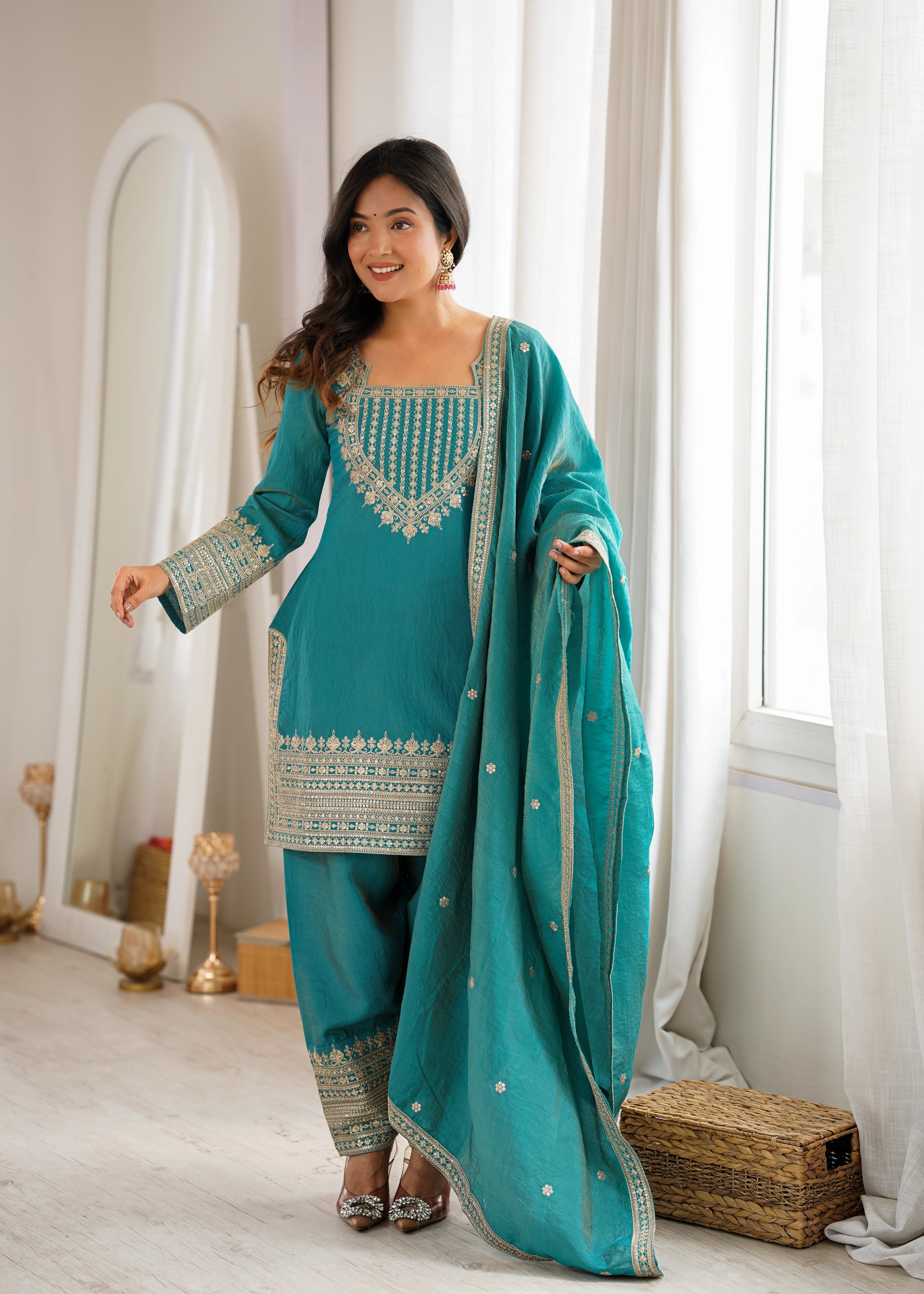 Woman in a teal traditional outfit standing in a bright room with large windows.
