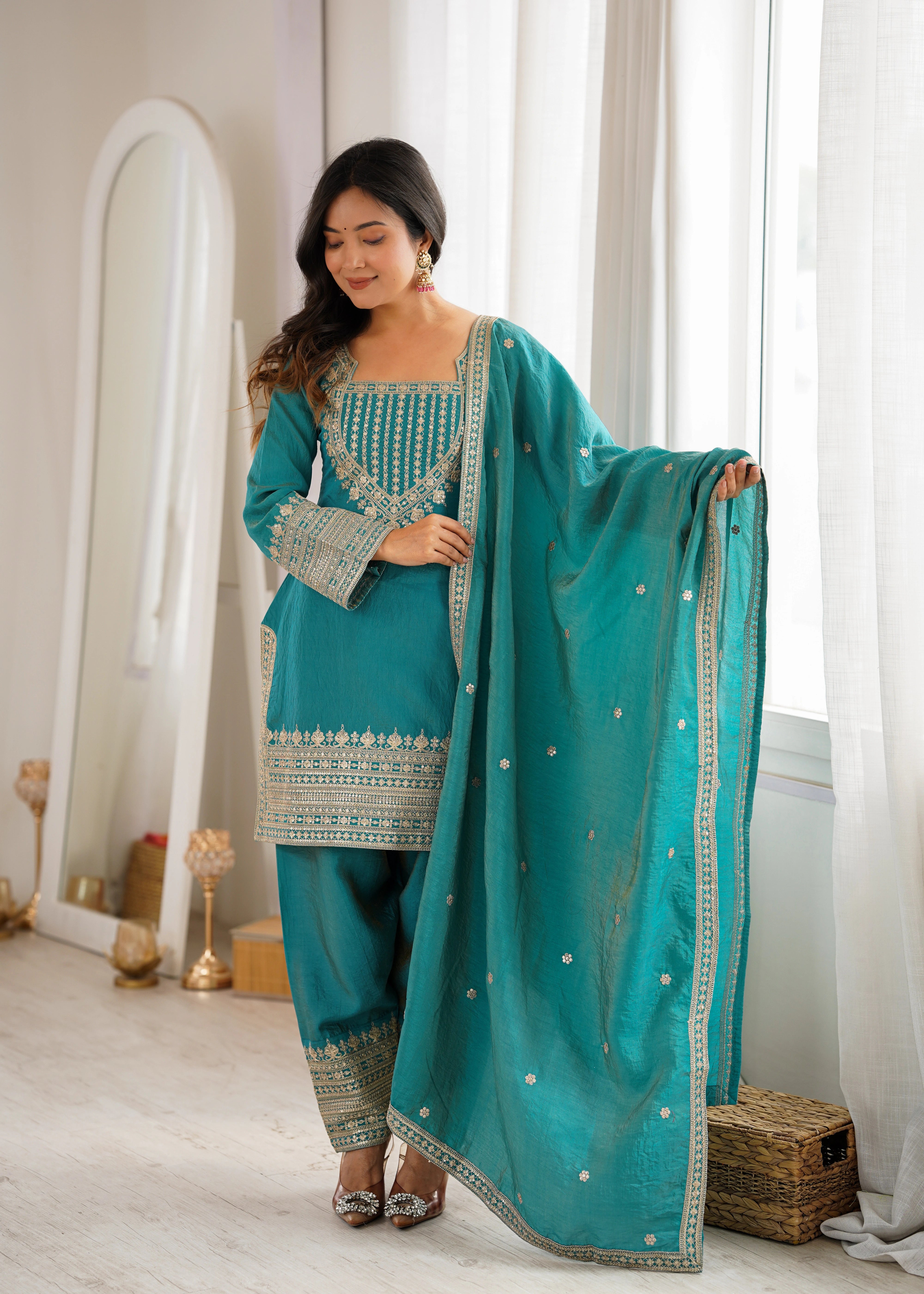 Woman in a teal traditional outfit with gold embroidery standing in a bright room.