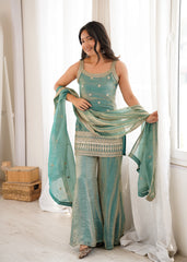 Woman in a teal traditional outfit with a matching dupatta, standing in a bright room.