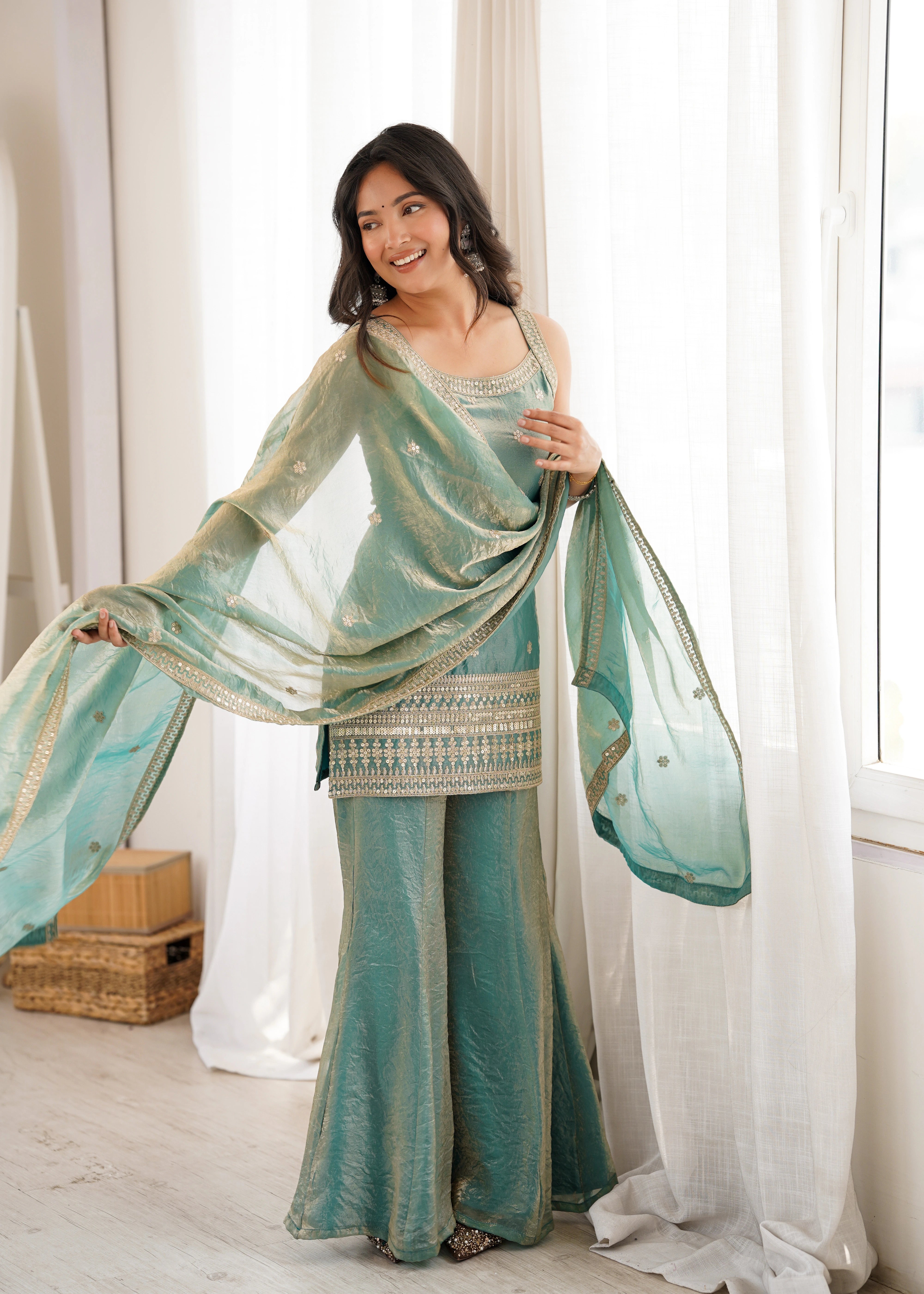 Woman in a teal saree with a matching blouse standing in a bright room with white curtains.