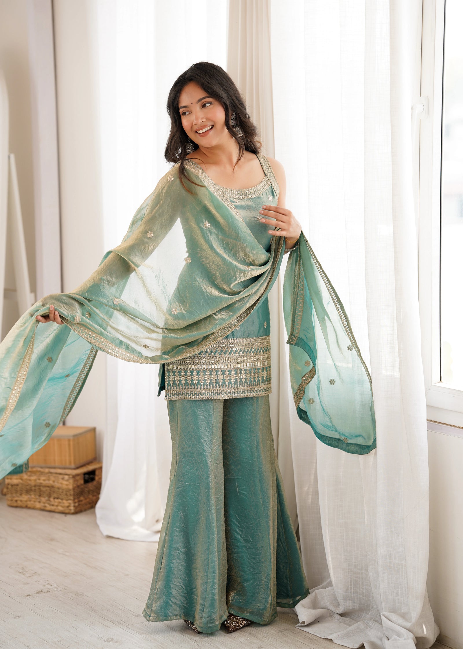 Woman in a teal saree with a matching blouse standing in a bright room with white curtains.