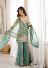 Woman in a teal saree with a matching blouse standing in a bright room with white curtains.