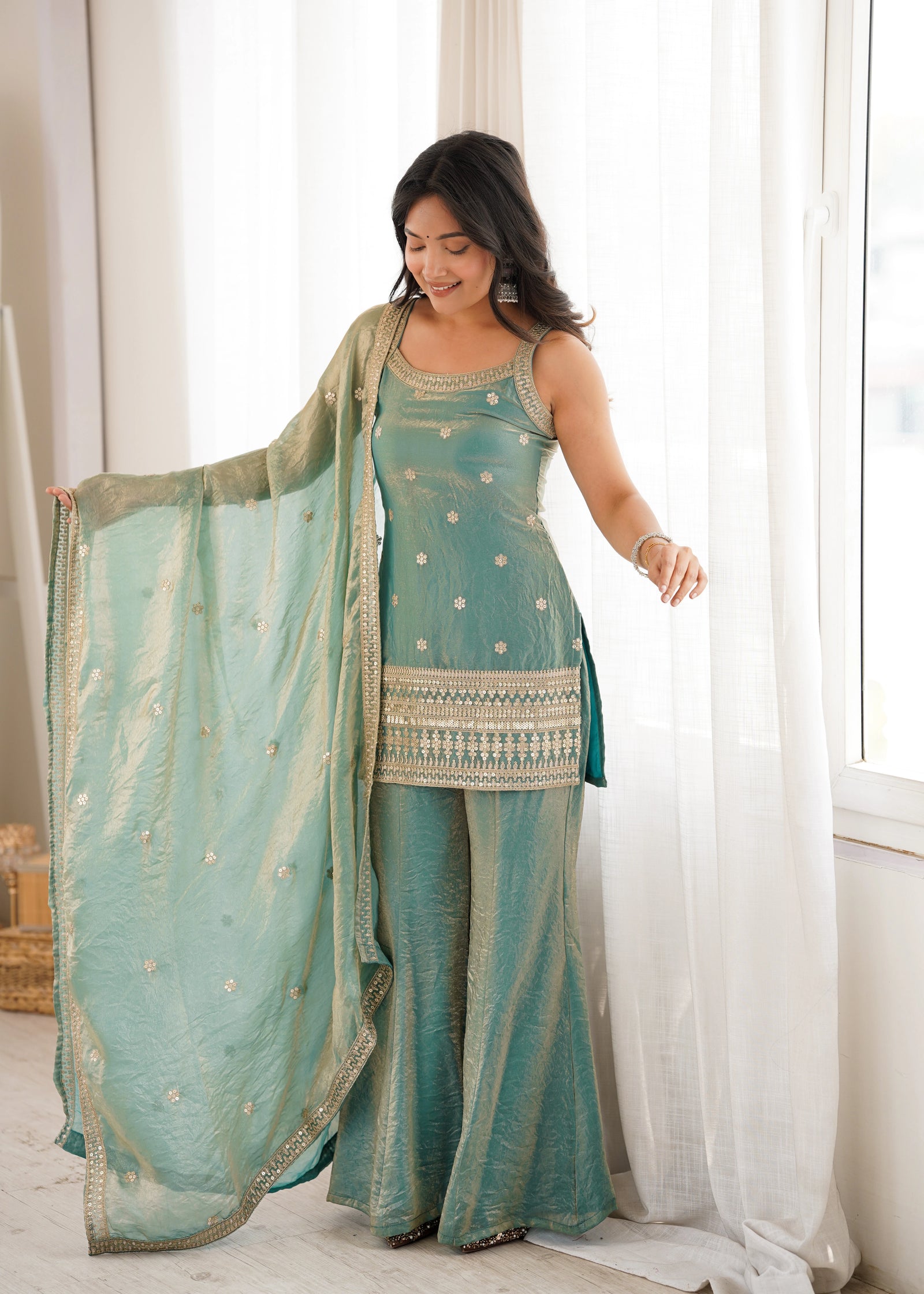 Woman in a traditional teal outfit with a matching dupatta standing in a bright room.