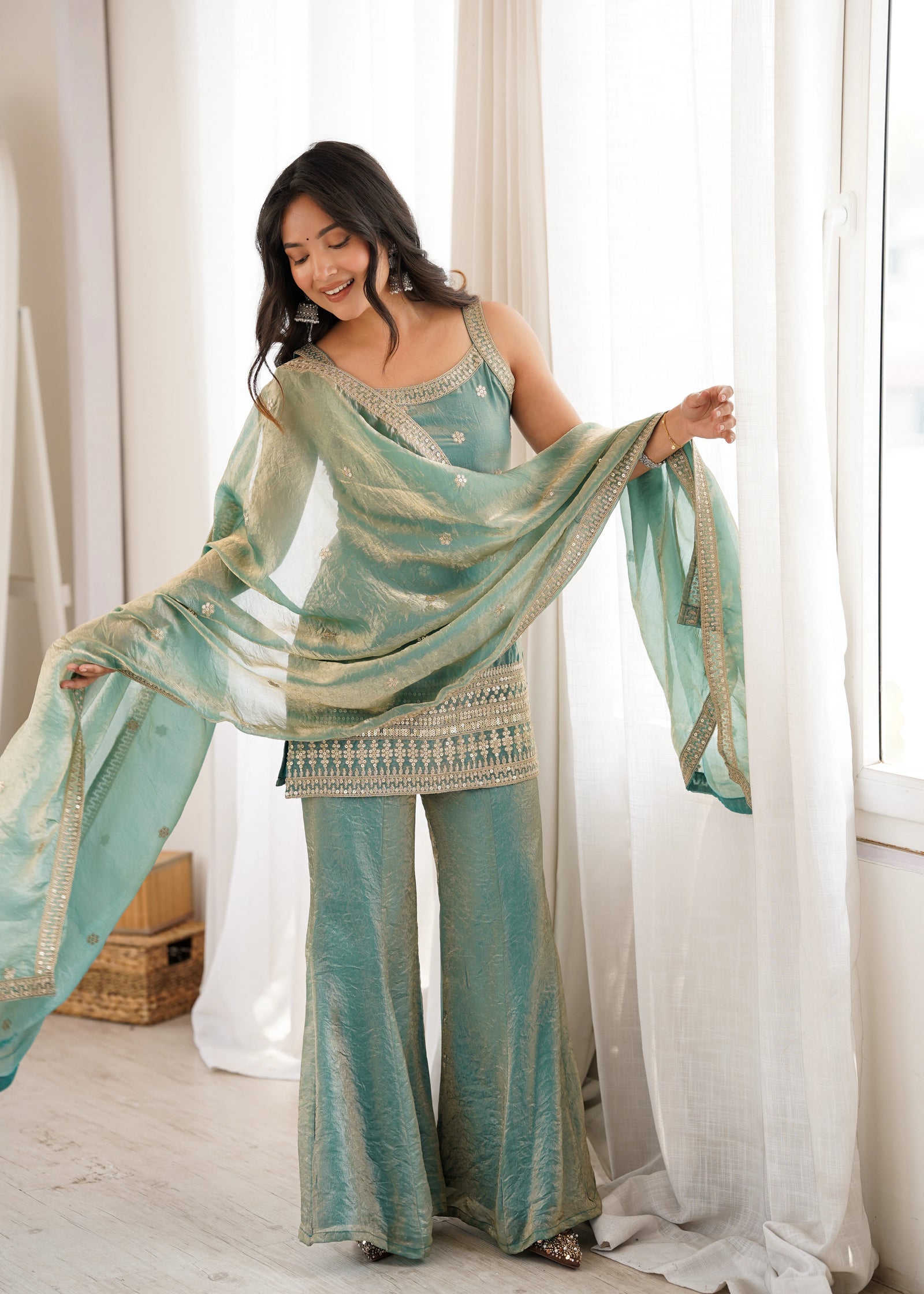 Woman wearing a teal and gold traditional outfit with a matching dupatta in a bright room.
