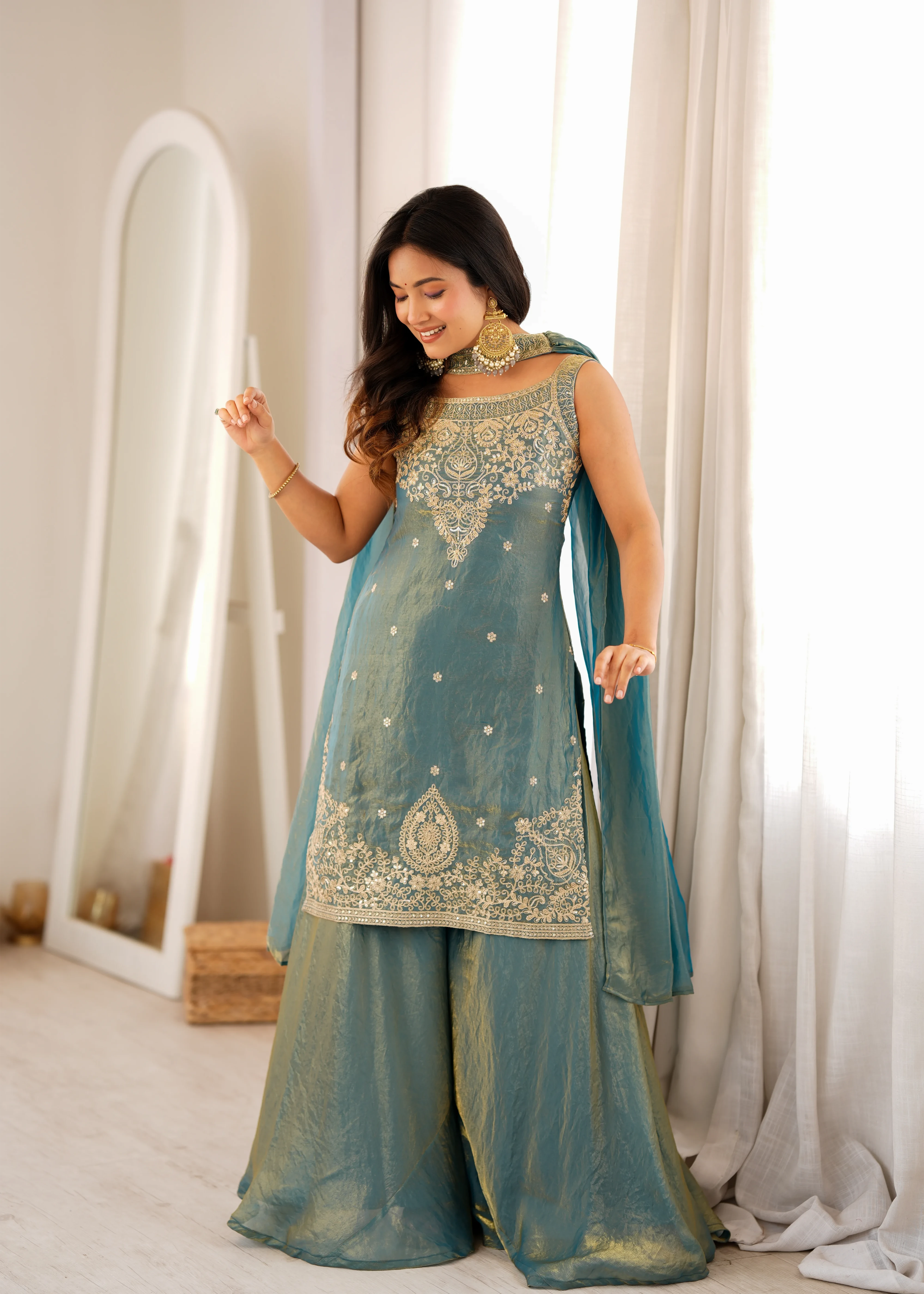 Woman in a teal traditional outfit with gold embroidery standing in a well-lit room.