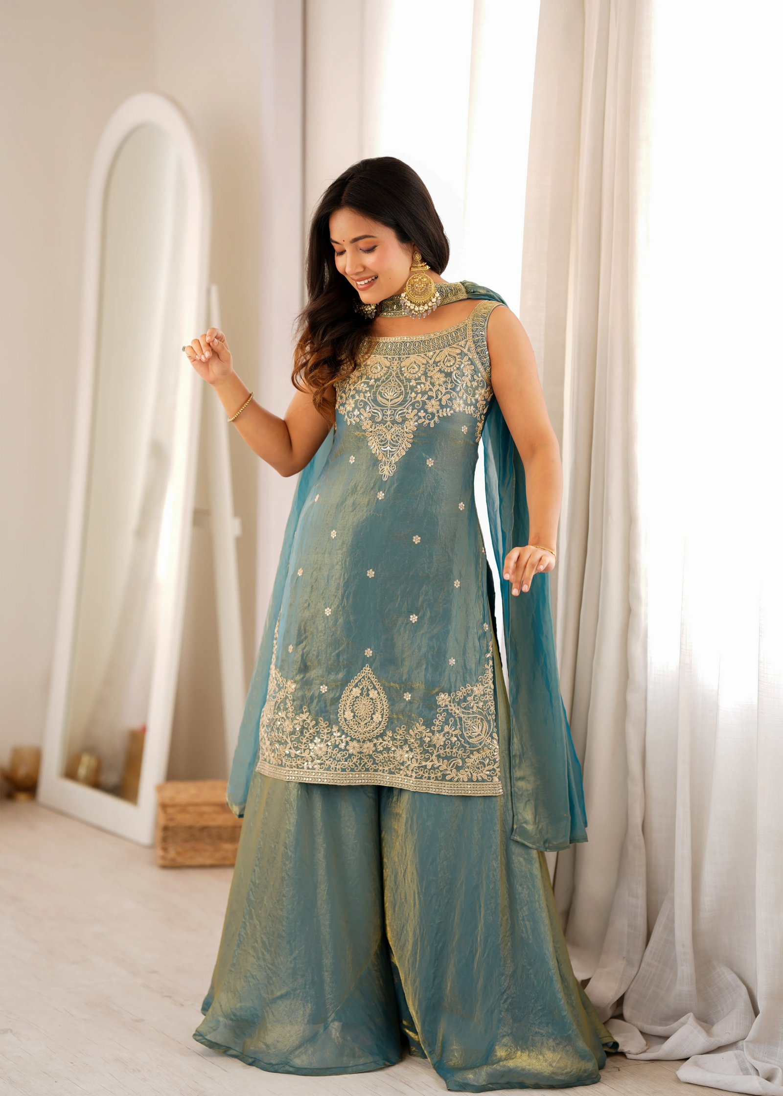 Woman in a teal traditional outfit with gold embroidery standing in a well-lit room.