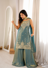 Woman in a teal traditional outfit with gold embroidery standing in a well-lit room.