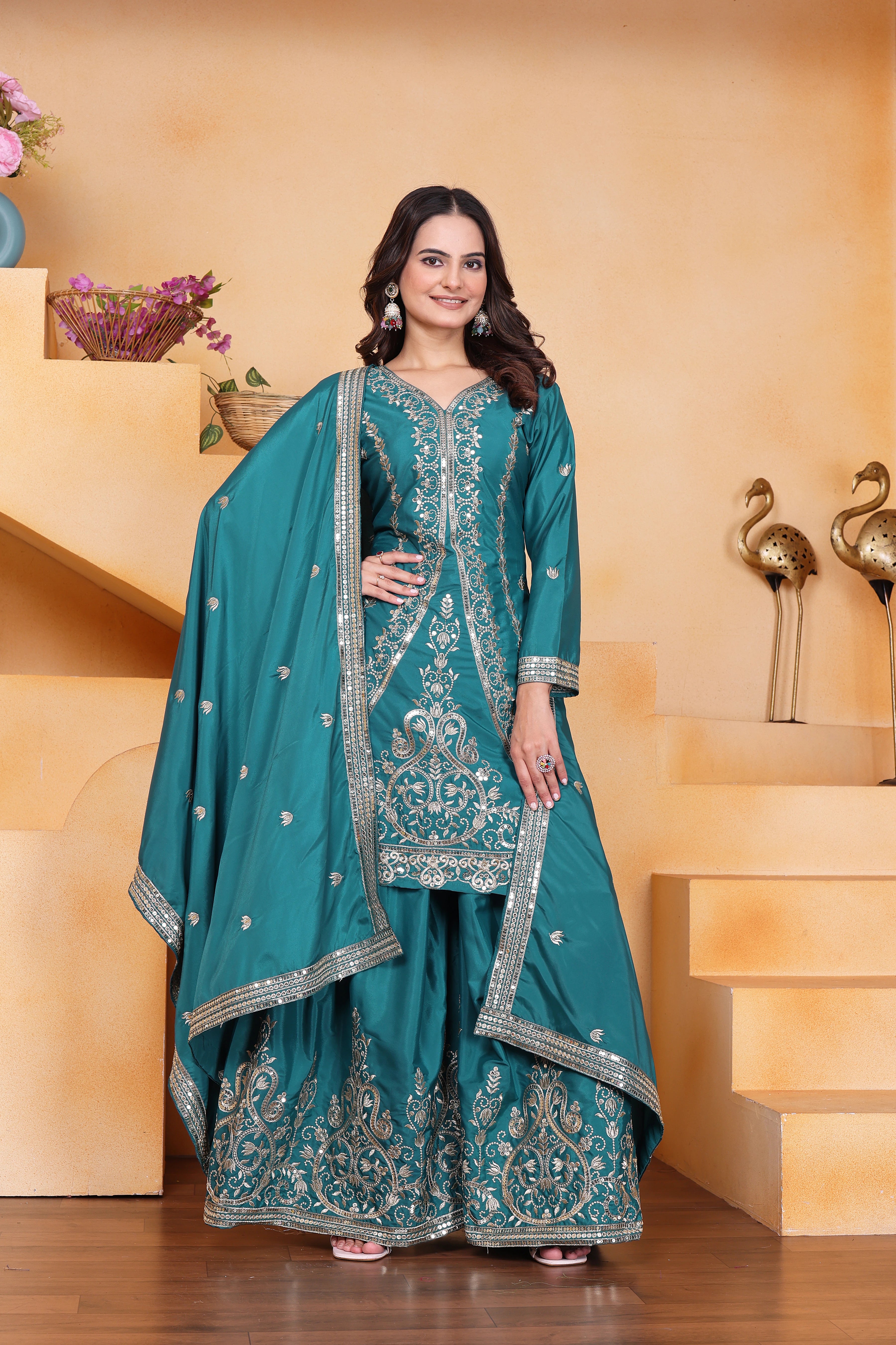 Woman in a teal traditional outfit with silver embroidery standing indoors.