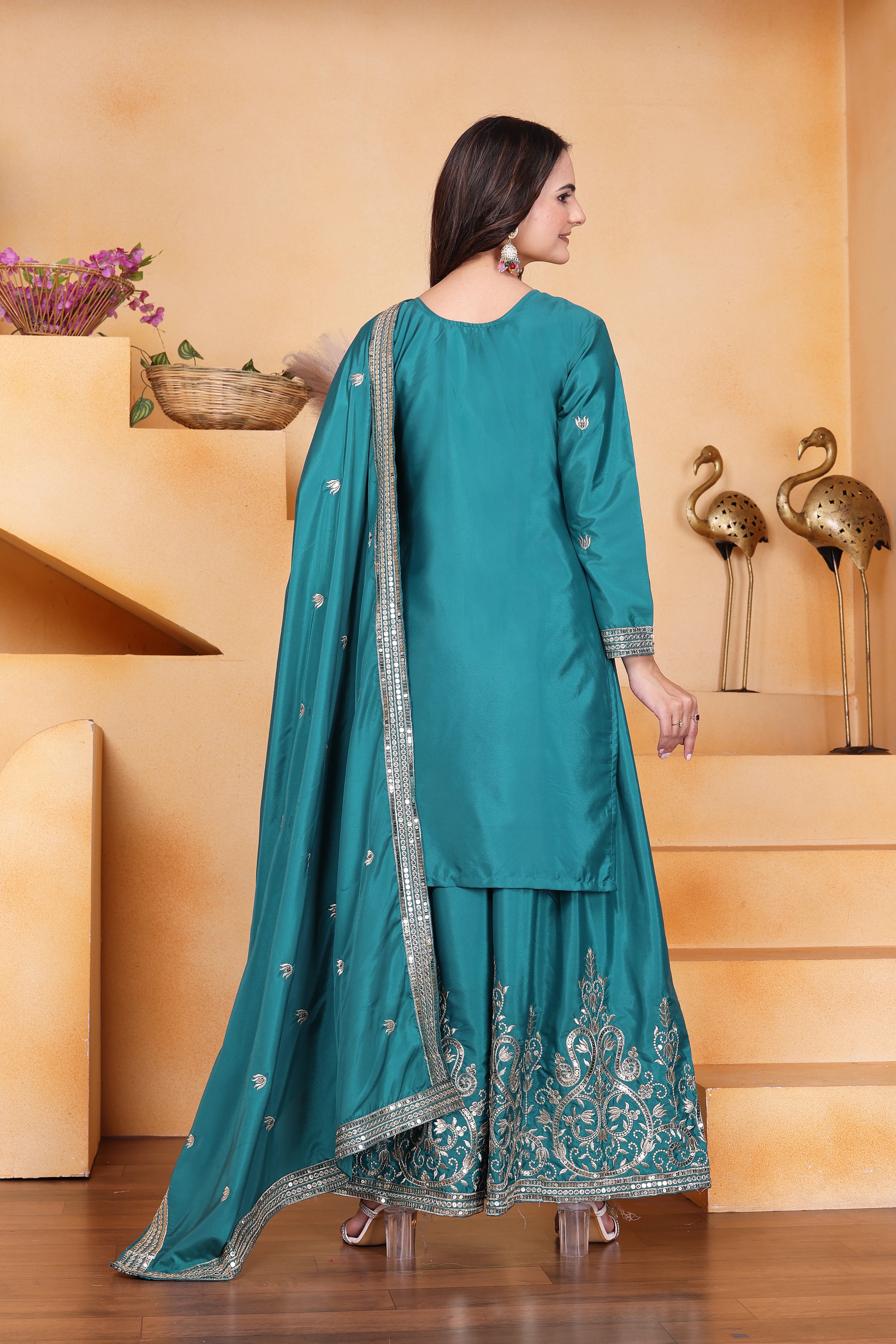 Woman in a teal traditional outfit with silver embroidery standing in a room with decorative elements.