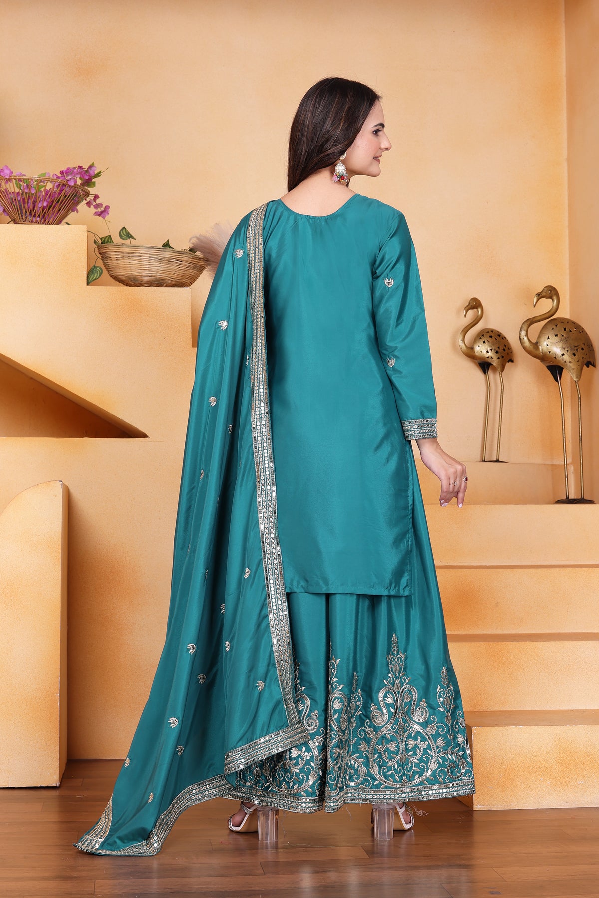 Woman in a teal traditional outfit with silver embroidery standing in a room with decorative elements.