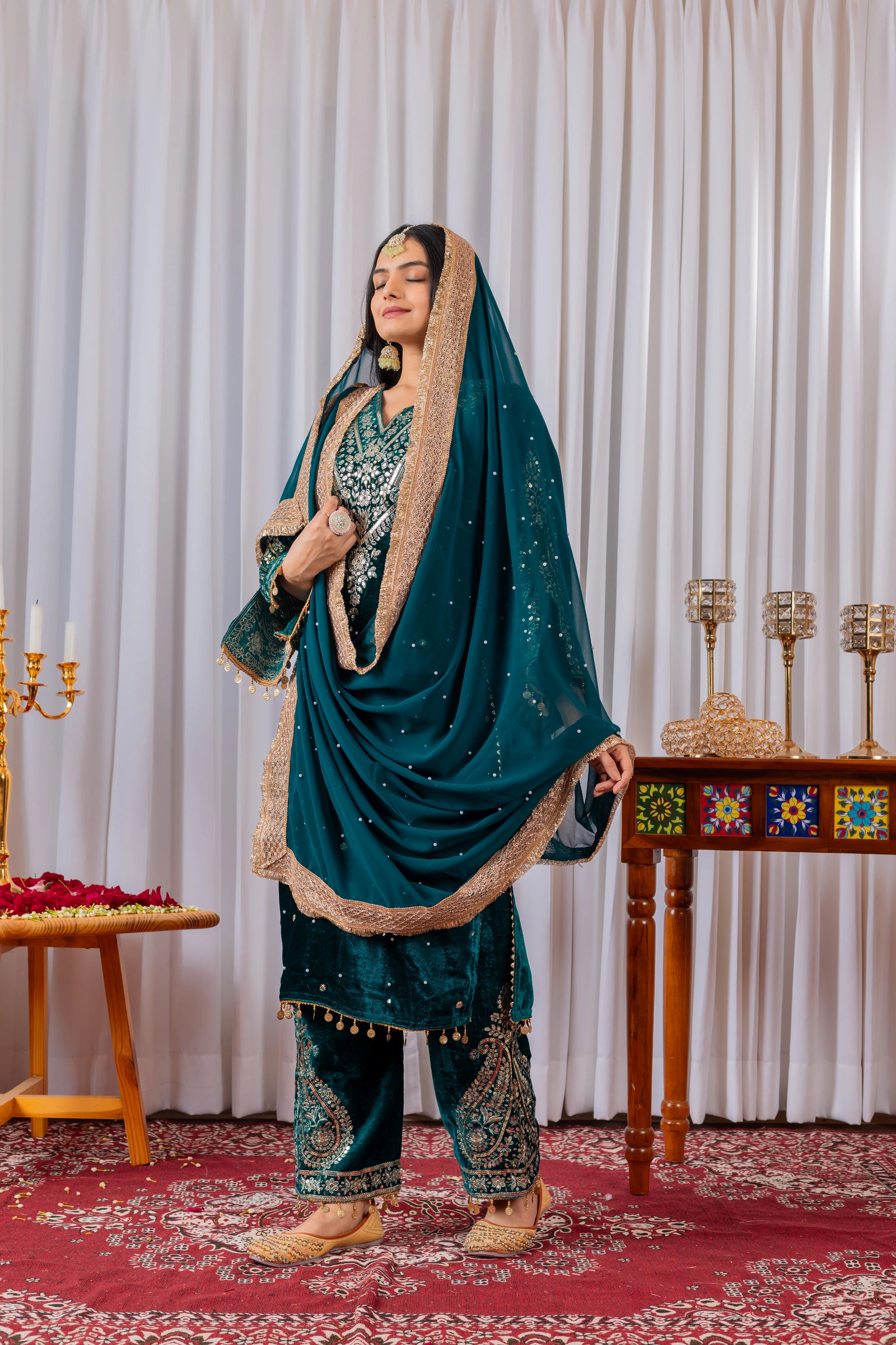 Woman in traditional teal and gold outfit standing in a decorated room with tables and candles.