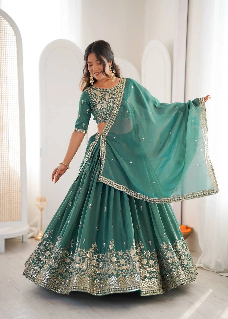 Woman in a teal embroidered traditional outfit standing in a bright room.
