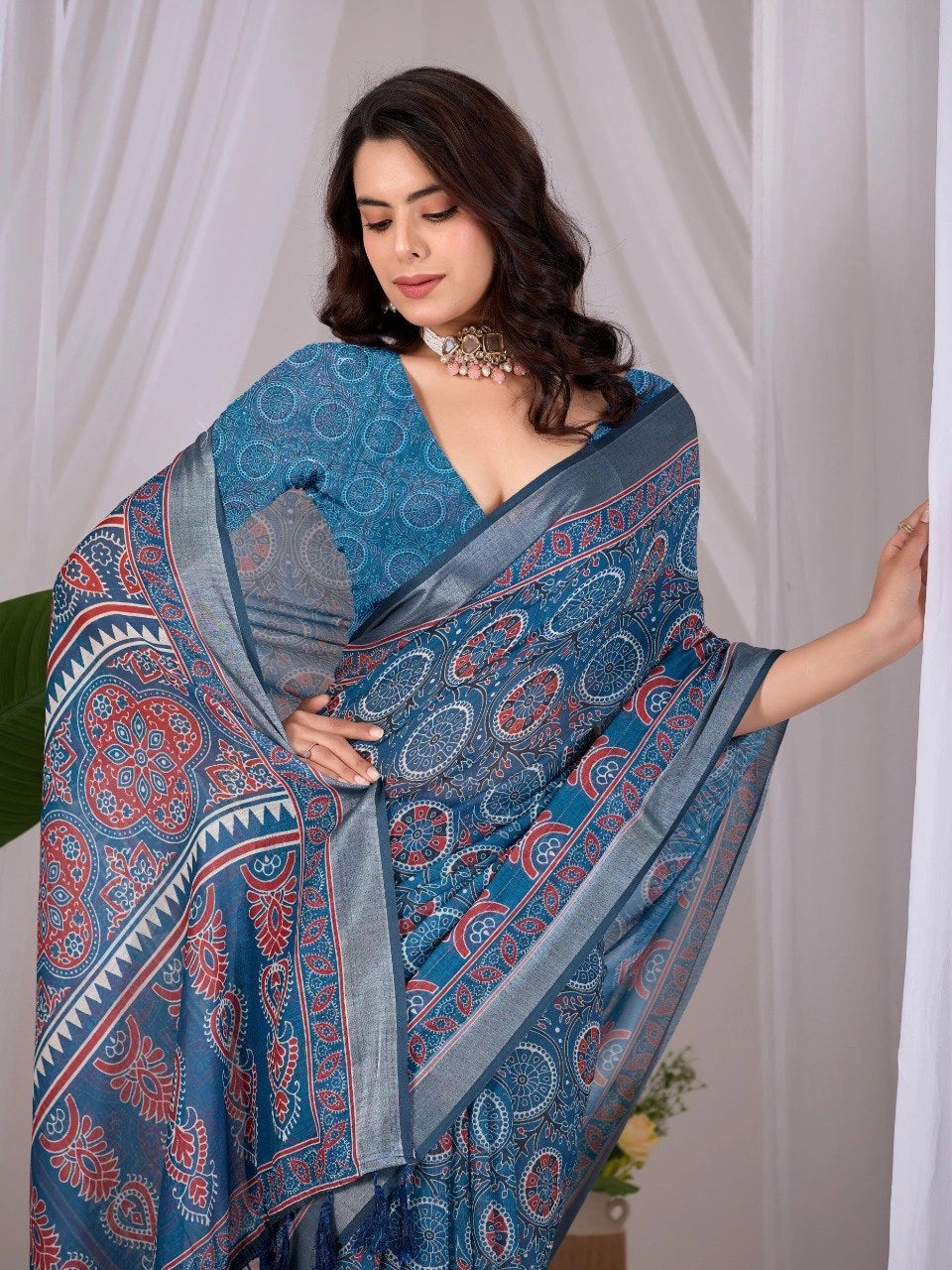 Woman wearing a blue saree with a pattern, standing against a white curtain.