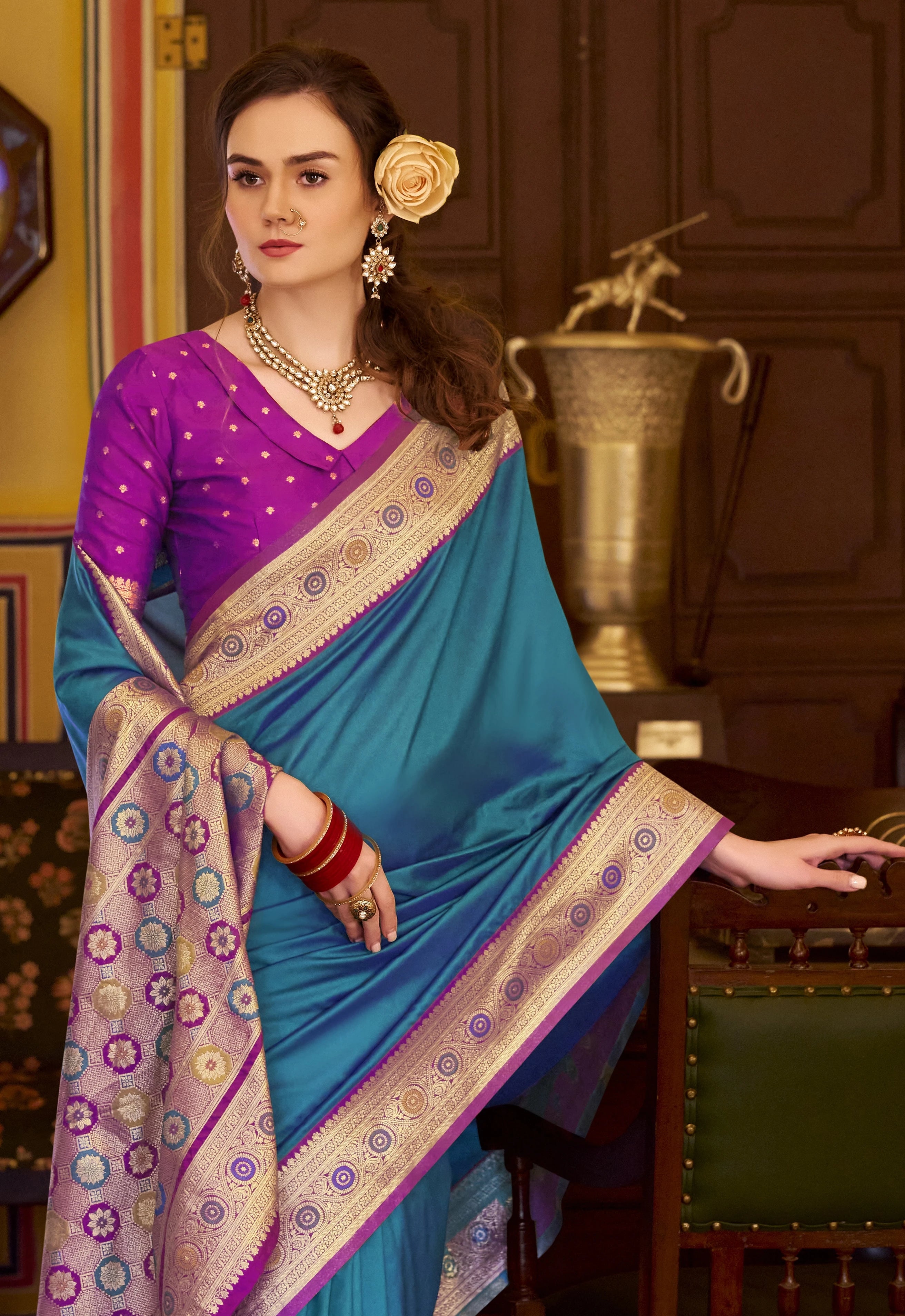 Woman wearing a blue and purple saree with a decorative flower in her hair, sitting in an elegant room.