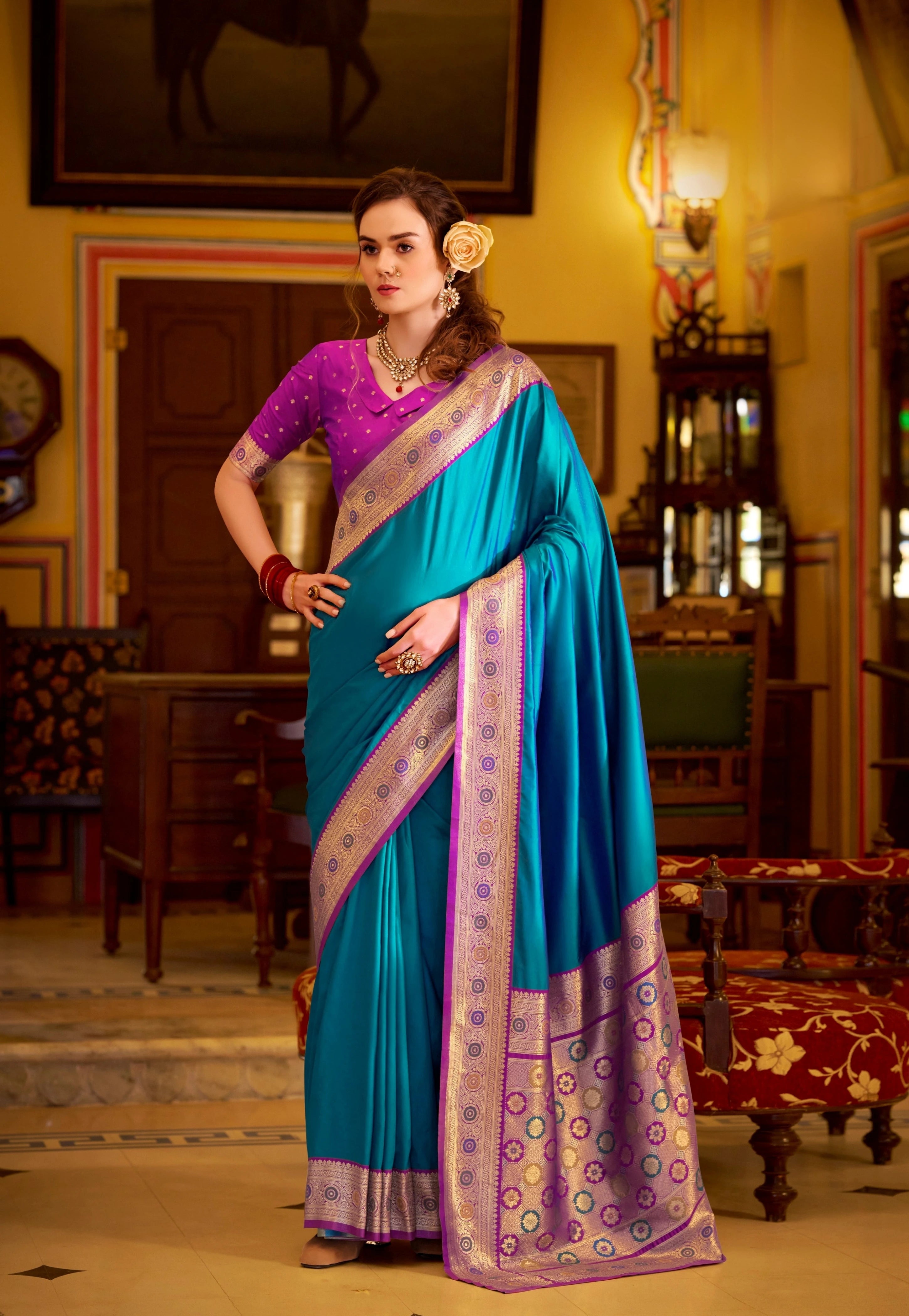 Woman in a blue and purple saree standing in an ornate room.