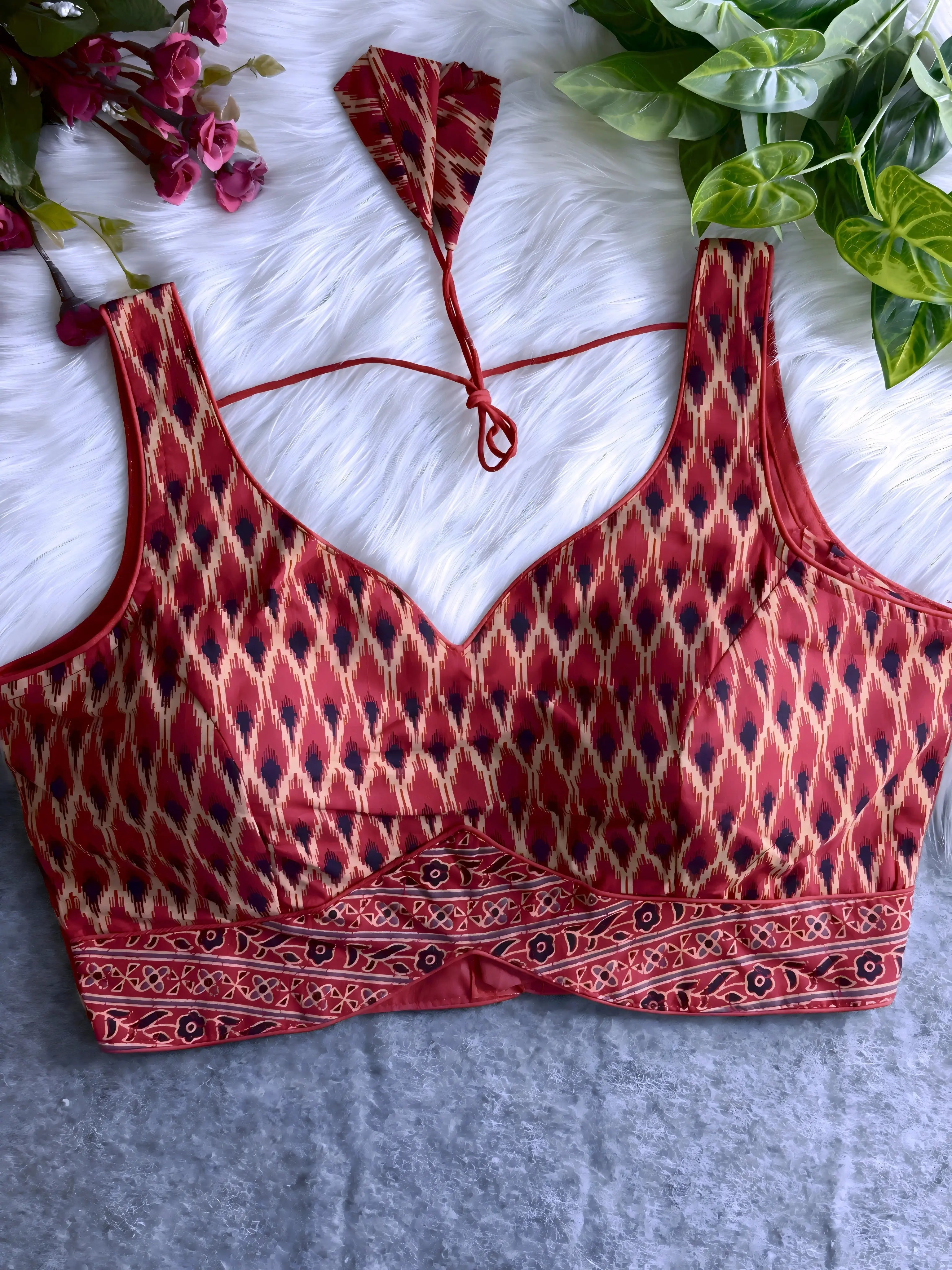 Maroon silk blouse featuring traditional Ikat-style geometric print in cream and black, wide shoulder straps, structured cups, and a patterned border at the hem. The design includes a V-neck and matching tasseled tie-back detail, displayed flat on a soft white fur background with artificial pink and green foliage arranged at the corners.