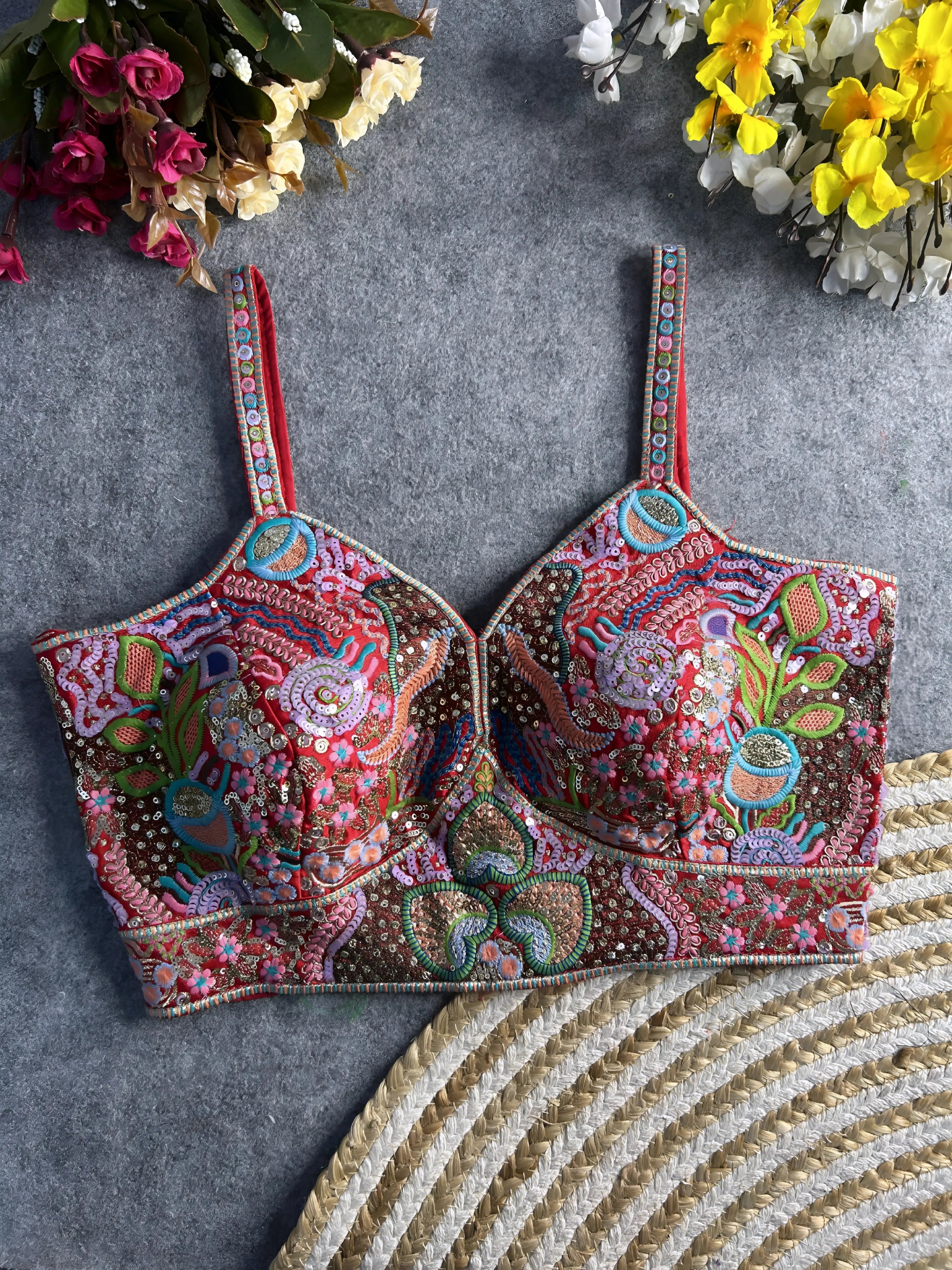Traditional multicolor bustier blouse featuring vibrant Madhubani-inspired embroidery with intricate floral, paisley, and geometric motifs, embellished with sequins and beads, structured cups, and adjustable straps, displayed flat on a textured grey surface with artificial pink, yellow, and white flowers in the corners.
