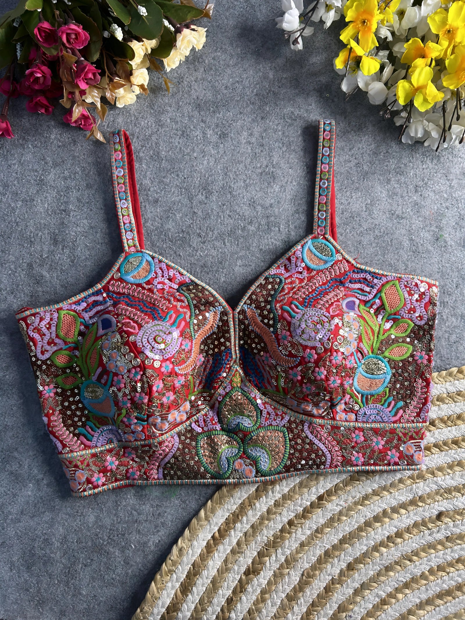 Traditional multicolor bustier blouse featuring vibrant Madhubani-inspired embroidery with intricate floral, paisley, and geometric motifs, embellished with sequins and beads, structured cups, and adjustable straps, displayed flat on a textured grey surface with artificial pink, yellow, and white flowers in the corners.