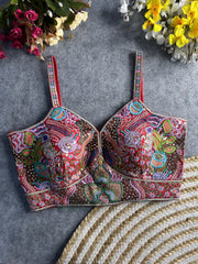 Traditional multicolor bustier blouse featuring vibrant Madhubani-inspired embroidery with intricate floral, paisley, and geometric motifs, embellished with sequins and beads, structured cups, and adjustable straps, displayed flat on a textured grey surface with artificial pink, yellow, and white flowers in the corners.