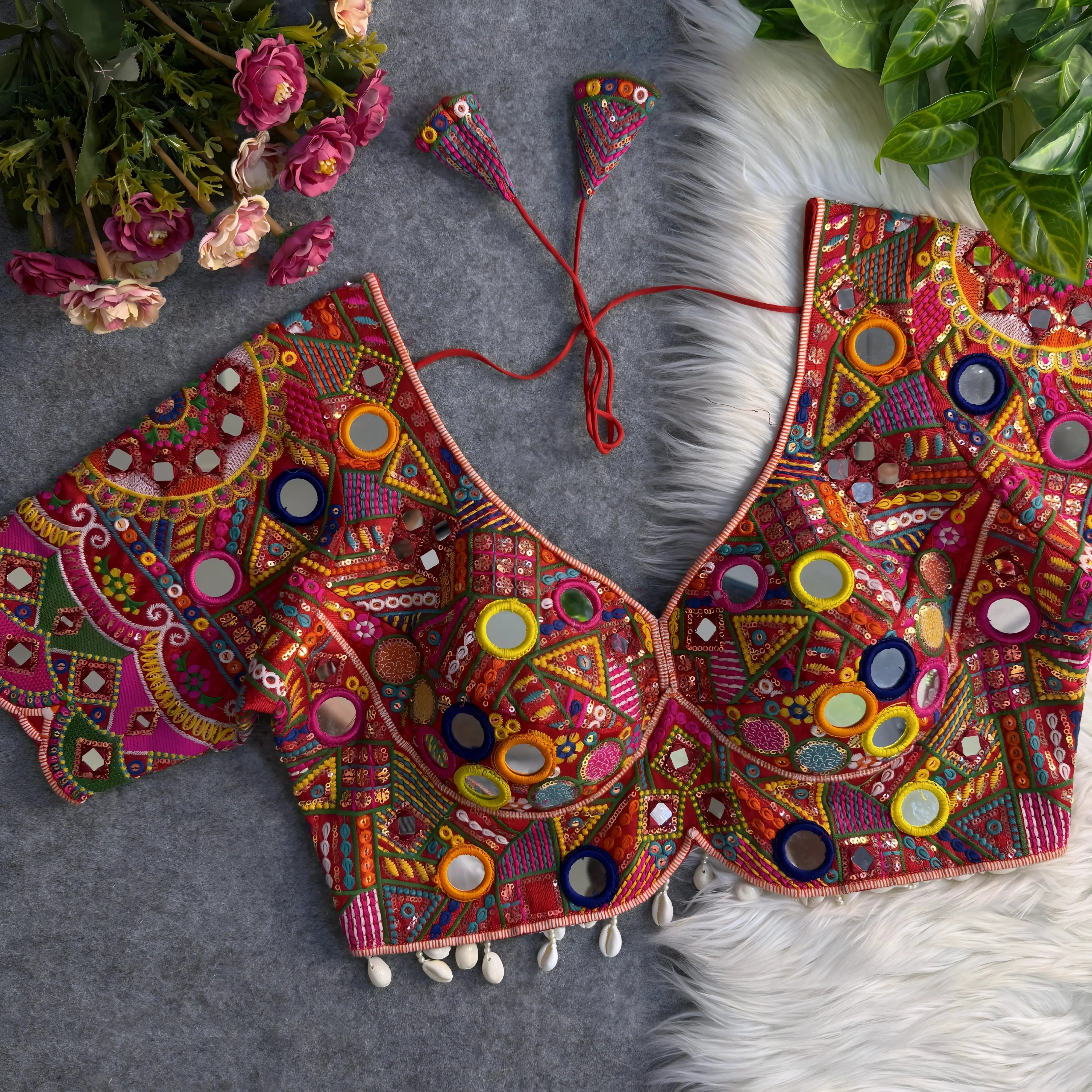 Traditional multicolor blouse featuring intricate Gujarati-style mirror embroidery with vibrant geometric and floral patterns in pink, yellow, orange, green, blue, and red. The blouse has wide short sleeves, circular and teardrop mirrors, shell-beaded trim at the hem, and tasseled tie-back detail. Displayed flat on a grey surface with artificial pink flowers and green foliage in the corners.