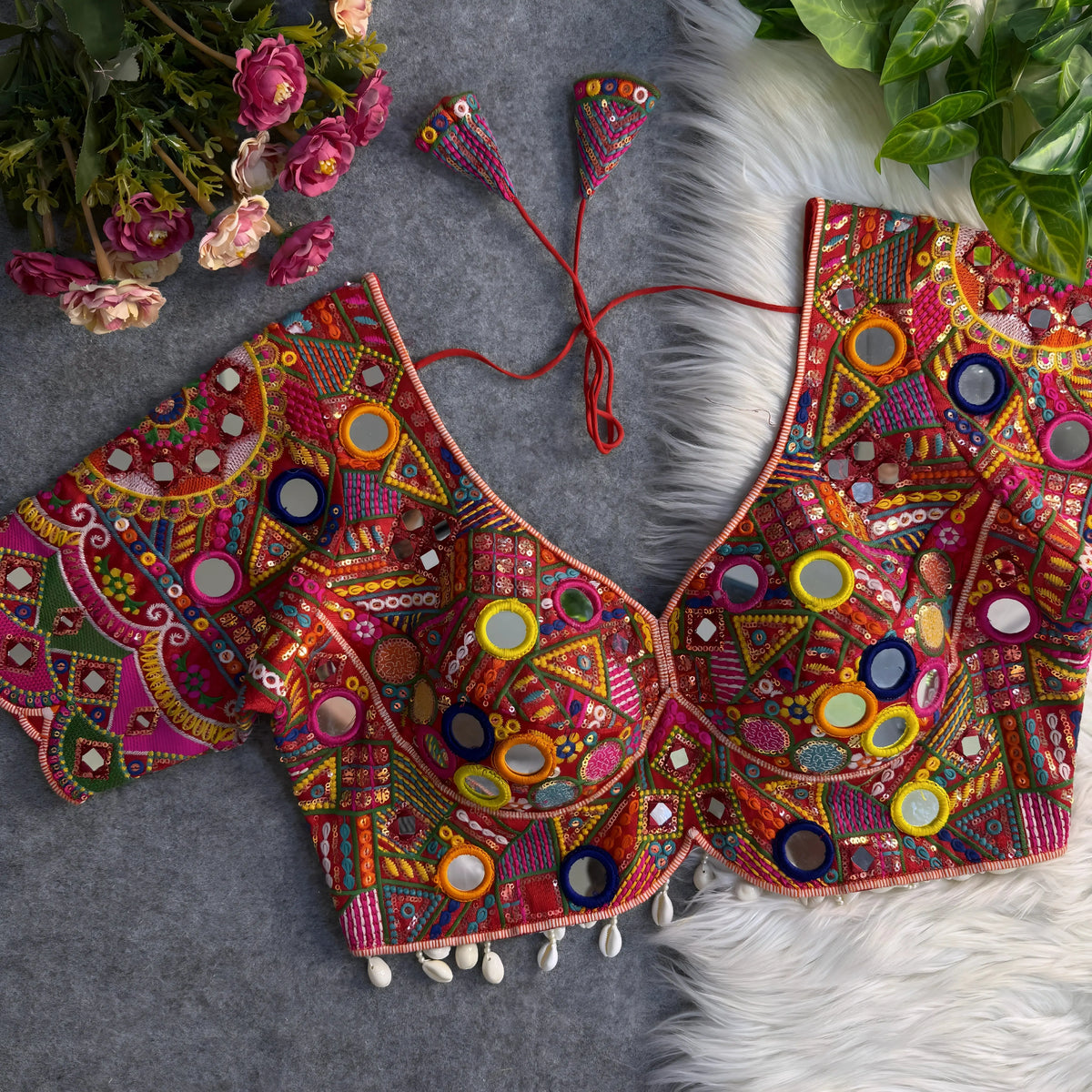 Traditional multicolor blouse featuring intricate Gujarati-style mirror embroidery with vibrant geometric and floral patterns in pink, yellow, orange, green, blue, and red. The blouse has wide short sleeves, circular and teardrop mirrors, shell-beaded trim at the hem, and tasseled tie-back detail. Displayed flat on a grey surface with artificial pink flowers and green foliage in the corners.