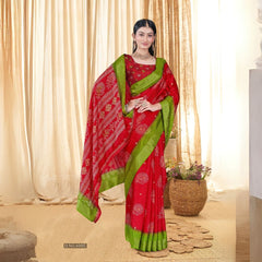 Woman in a red sequence jacquard work saree with green border standing indoors against a backdrop of beige curtains and woven decor elements